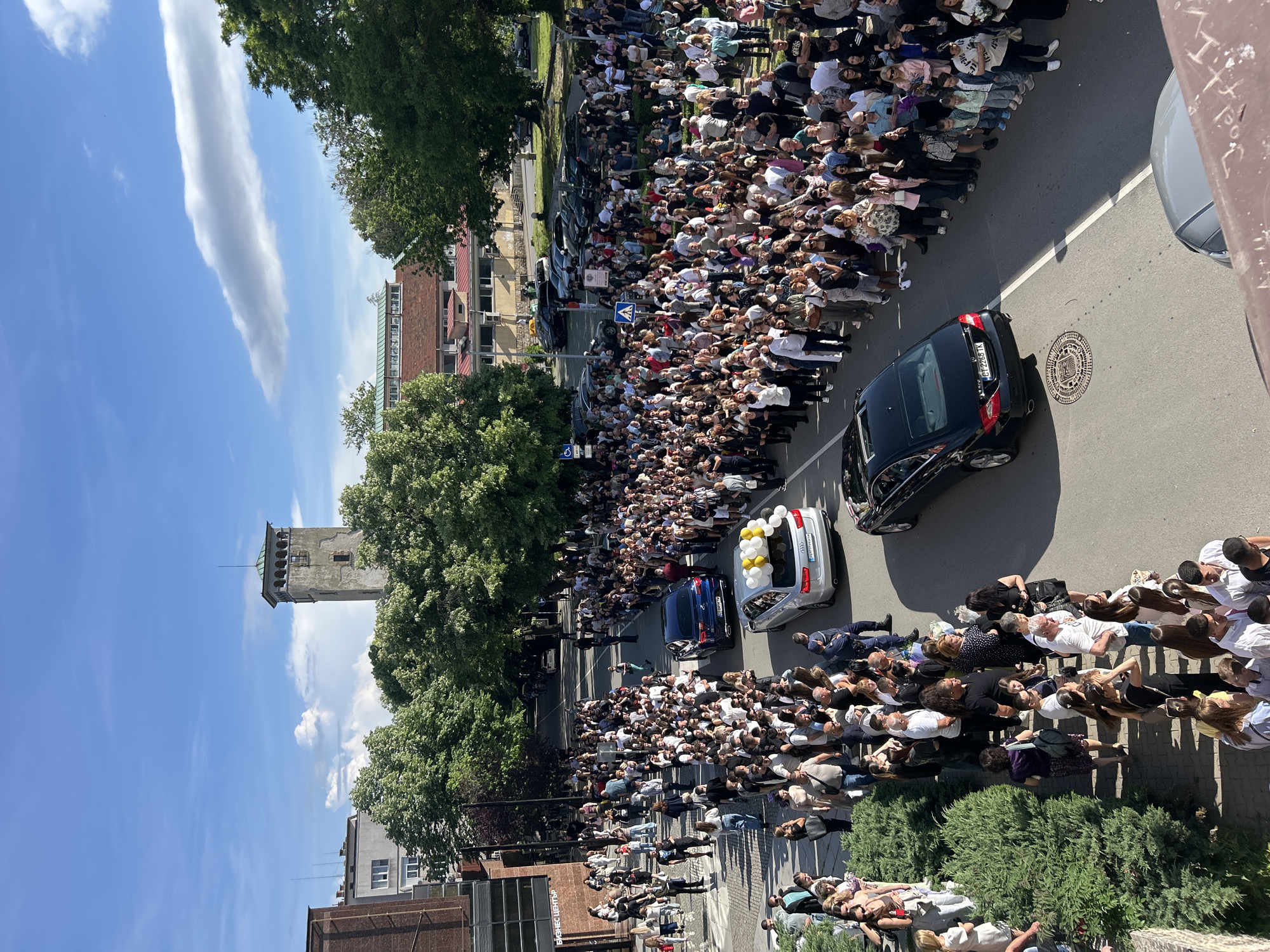 The image captures a street scene in an urban environment. A large crowd of people lines both sides of the street, forming a dense pathway. The majority of the people appear to be civilians of diverse ages and appearances. Three vehicles, a black car, a blue car, and a silver car with balloons attached, are navigating the street. The presence of balloons suggests a celebratory or commemorative event. The street itself is paved and includes a manhole cover.
Buildings, trees, and a tower structure are visible in the background. The tower is tall, and it appears to be an old structure. Trees are abundant along the road. The sky is clear, with a few clouds. The presence of the crowd and vehicles indicates an event of significance is taking place in the street.