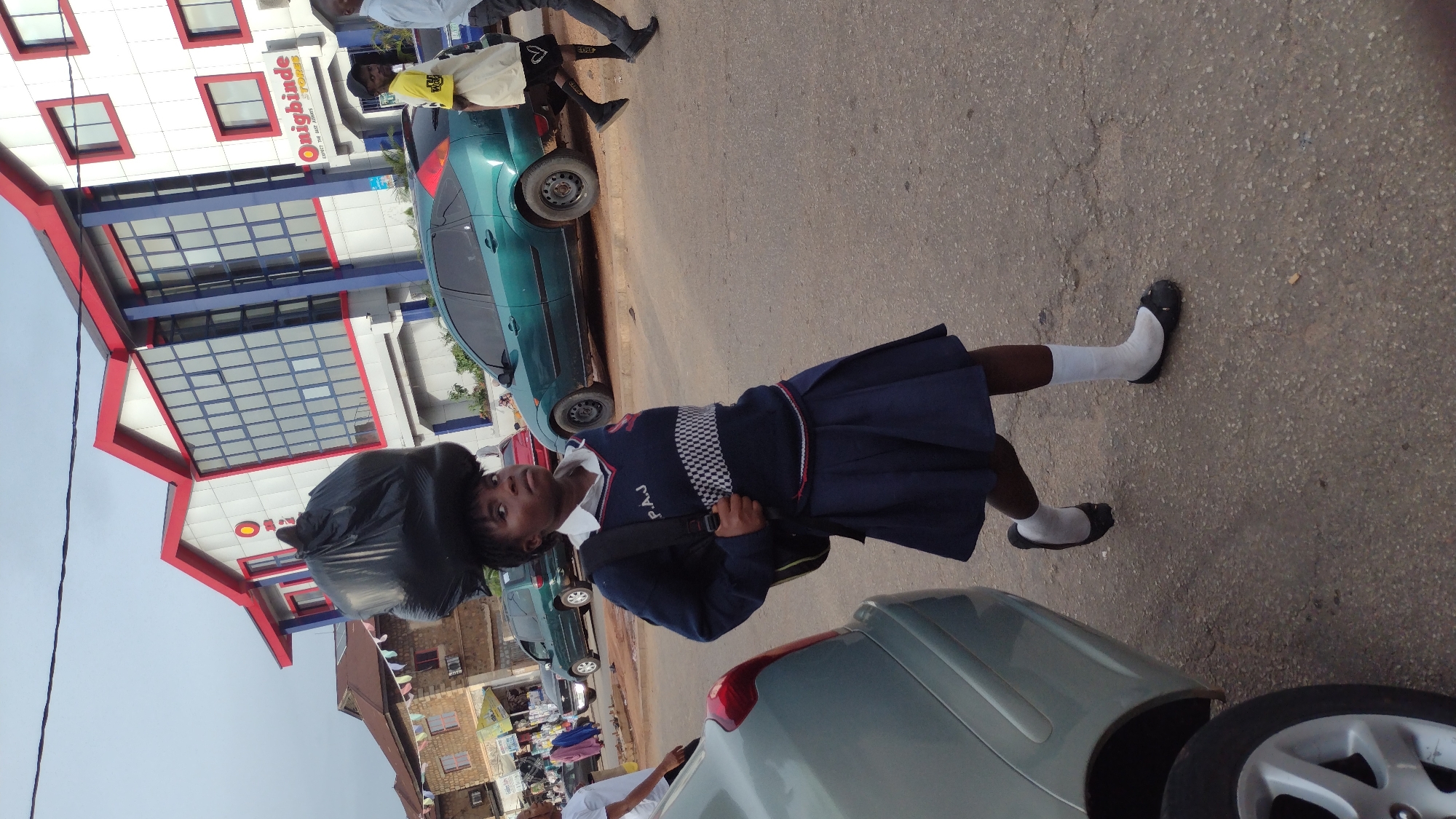 A young girl with a large black trash bag balanced on her head is walking on a paved road in Jos, Nigeria, near a modern building with red trim and large glass windows. She is wearing a dark blue school uniform with a white collared shirt, a V-neck sweater with a checkered pattern and "P.A.J." written on it, and a pleated skirt. She also has on white socks and black flat shoes. She is carrying a black backpack. Behind her, a teal hatchback car is parked on the side of the road, and other vehicles and buildings are visible in the background. The sky is overcast.