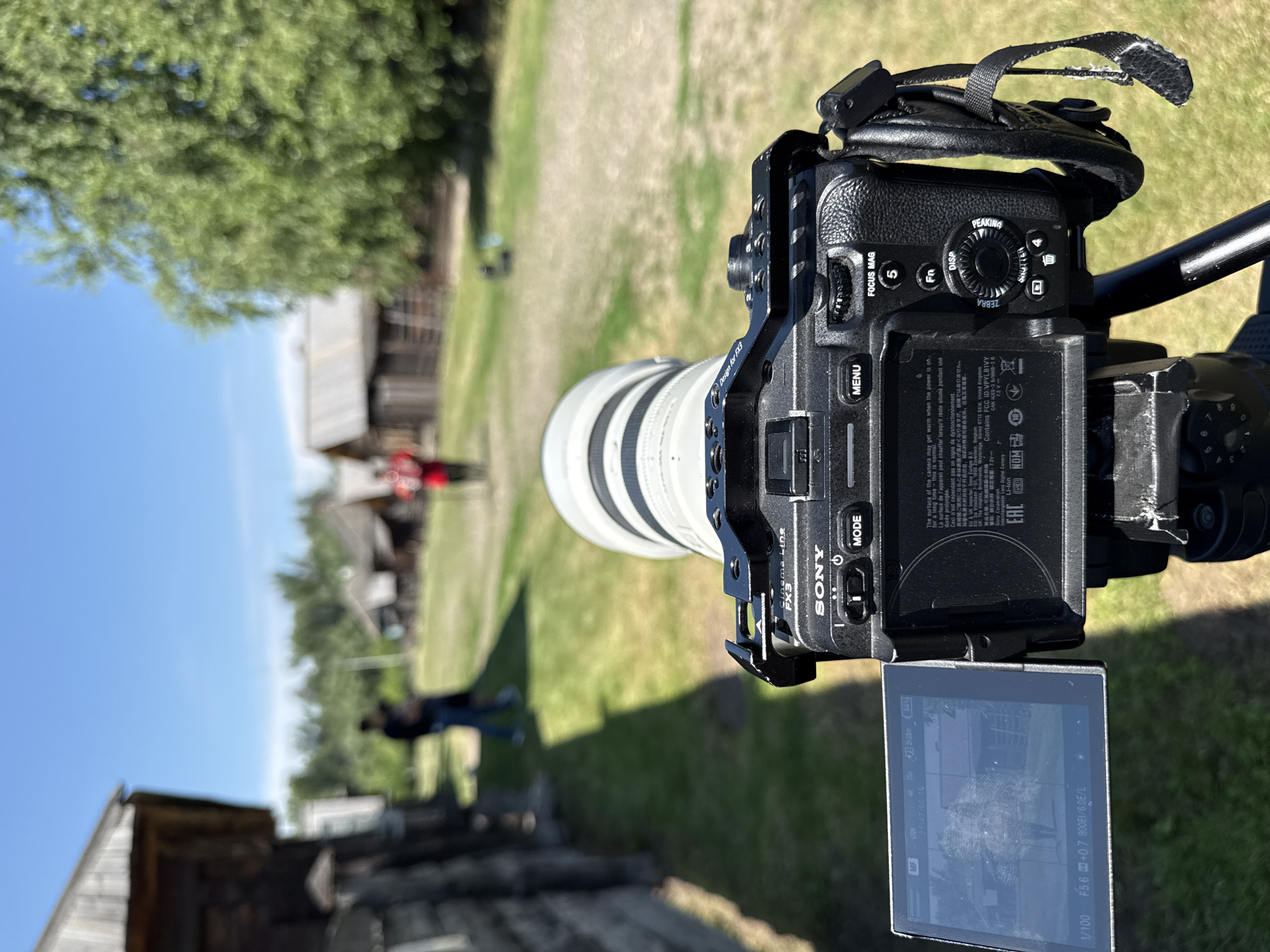 The image shows a Sony FX3 Cinema Line camera with a telephoto lens mounted on a tripod. The camera is aimed at a village scene with traditional wooden houses and a grassy path. In the background, two people are walking away from the camera, and another person in a red shirt is visible further down the path. The environment appears to be outdoors on a sunny day. The location is identified as Shushenskoye, Russia. The camera's LCD screen shows a preview of the scene.