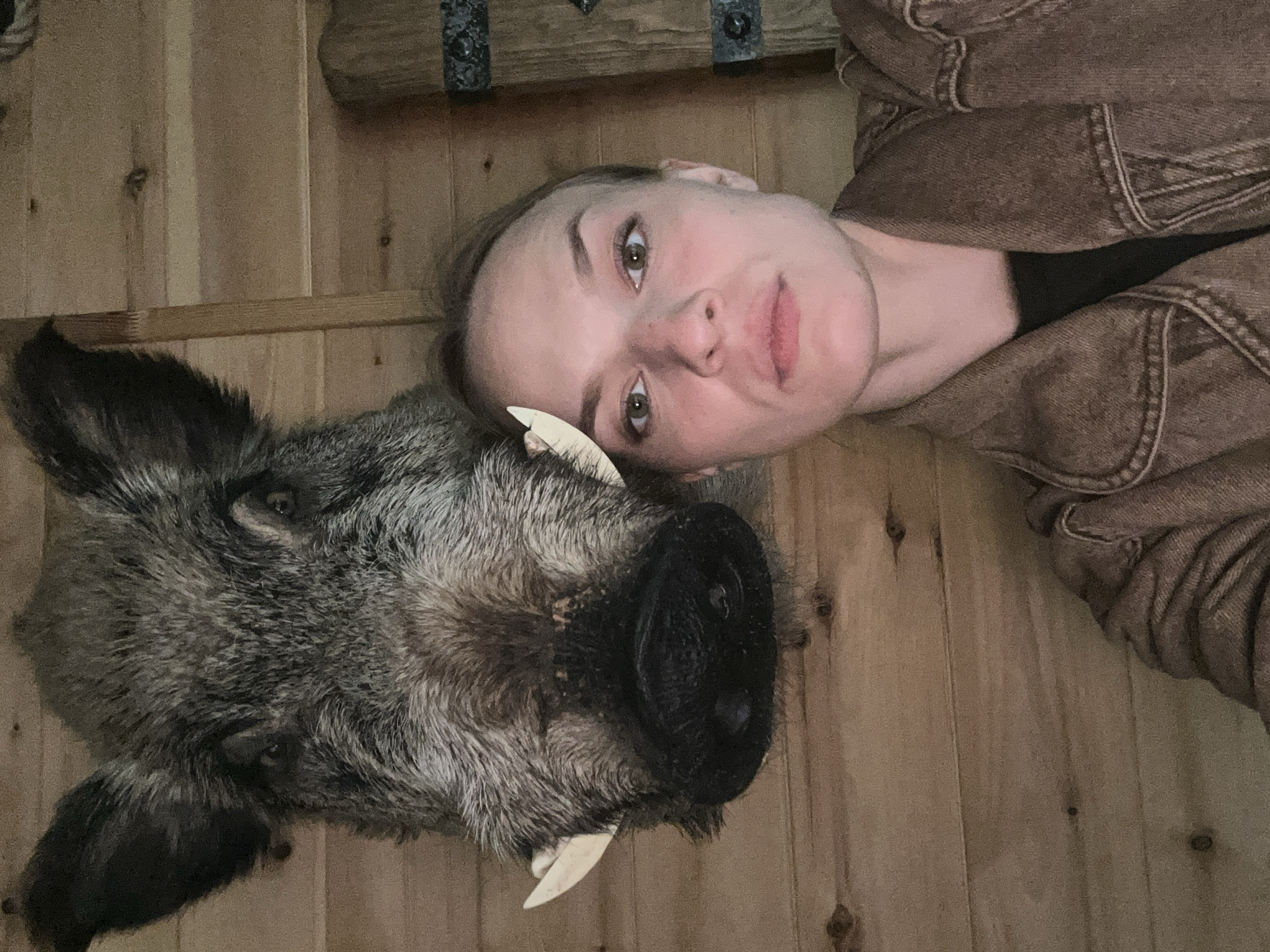 A woman with light skin, brown hair pulled back, and green eyes is taking a selfie in front of a taxidermied wild boar head. She is wearing a brown denim jacket over a dark shirt. The boar's head is mounted on a wooden wall, and its tusks are visible. The background consists of wooden plank walls, with a glimpse of what appears to be a decorative wooden object with metal accents on the right. The lighting is dim, casting shadows. There is no indication of the city of Karsha, Russia, in the image.