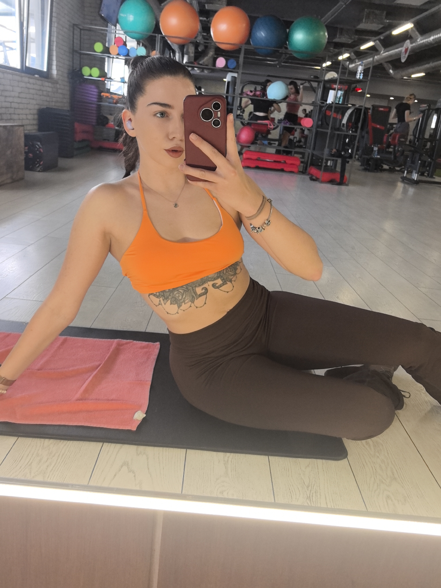 The image shows a young woman taking a selfie indoors, likely at a gym. She is sitting on a black yoga mat, wearing an orange sports bra and brown leggings. A pink towel lies to her left. She is holding a smartphone with a red case in front of her face. Her hair is pulled back in a ponytail. In the background, gym equipment and other people working out can be seen. The room is well-lit and appears to be a fitness center. While the city of Stara Zagora, Bulgaria is claimed, there is nothing in the image that confirms this.