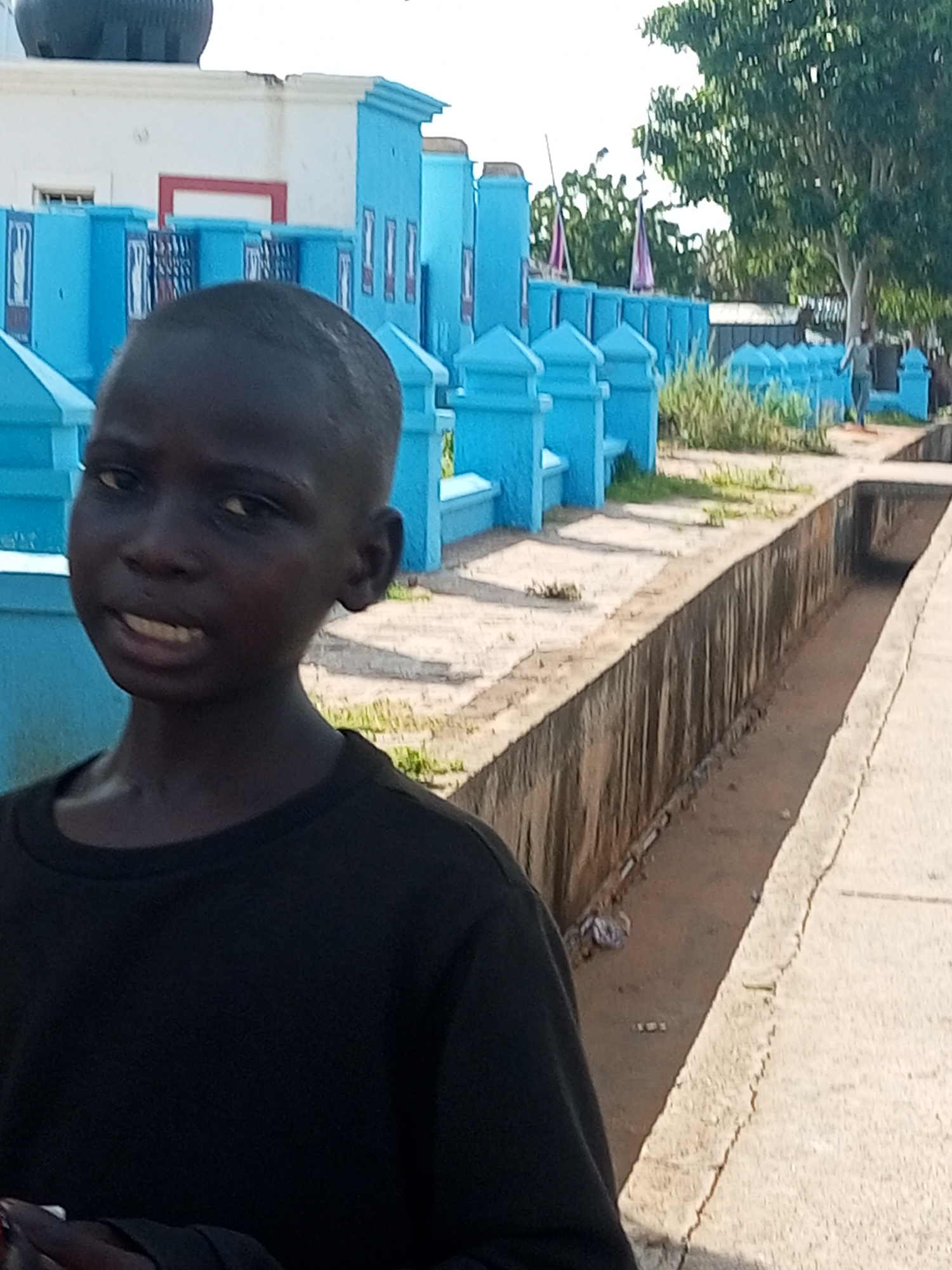 In Tempure, Nigeria, a young boy with dark skin and short hair stands in the foreground, wearing a black t-shirt. He is looking directly at the camera with his mouth slightly open. Behind him, a row of bright blue structures, possibly a cemetery or religious site, stretches into the distance. These structures are adorned with decorative elements and appear to be made of painted concrete. Beyond the blue structures, lush green trees are visible, and a person can be seen walking in the background. The ground in front of the blue structures is a mixture of dirt and paved areas, with a narrow, muddy trench running alongside a concrete pathway in the middle ground. The overall scene suggests an outdoor location in a moderately populated area, likely during daylight.