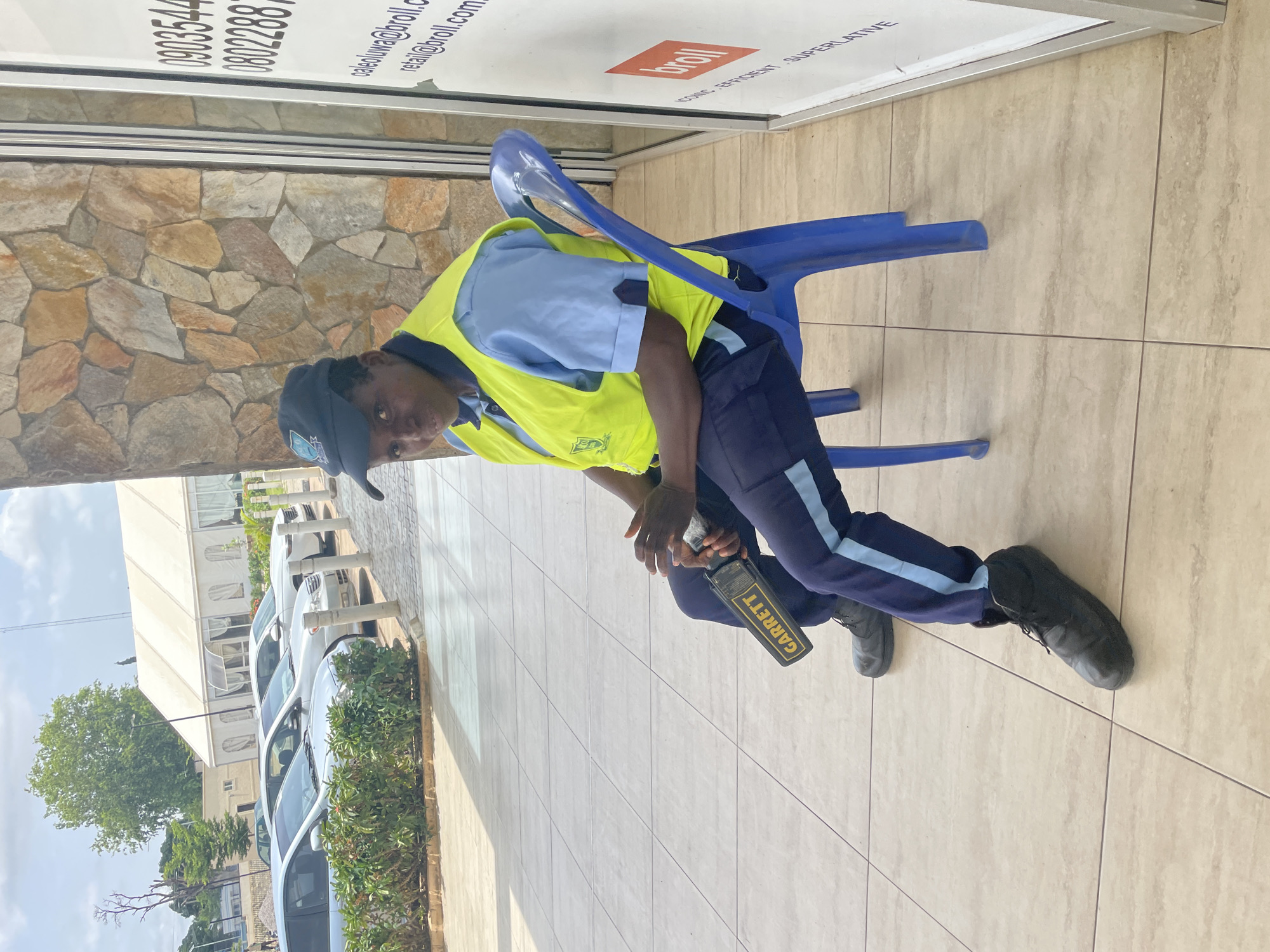 The image depicts a security guard seated on a blue plastic chair outdoors, possibly in the city of Unguwar Rimi, Nigeria. The guard, wearing a dark baseball cap, a light blue shirt, a neon yellow safety vest, and dark blue trousers with a light blue stripe, holds a black and yellow Garrett metal detector in their lap. The guard's left hand rests on the metal detector. The background shows a wall constructed of irregularly shaped stones of various earth tones, and to the right, a white vertical sign with text and a red rectangle. In the distance, several cars are parked in front of a large white tent. The ground is paved with beige tiles. The lighting suggests it is daytime with some cloud cover.