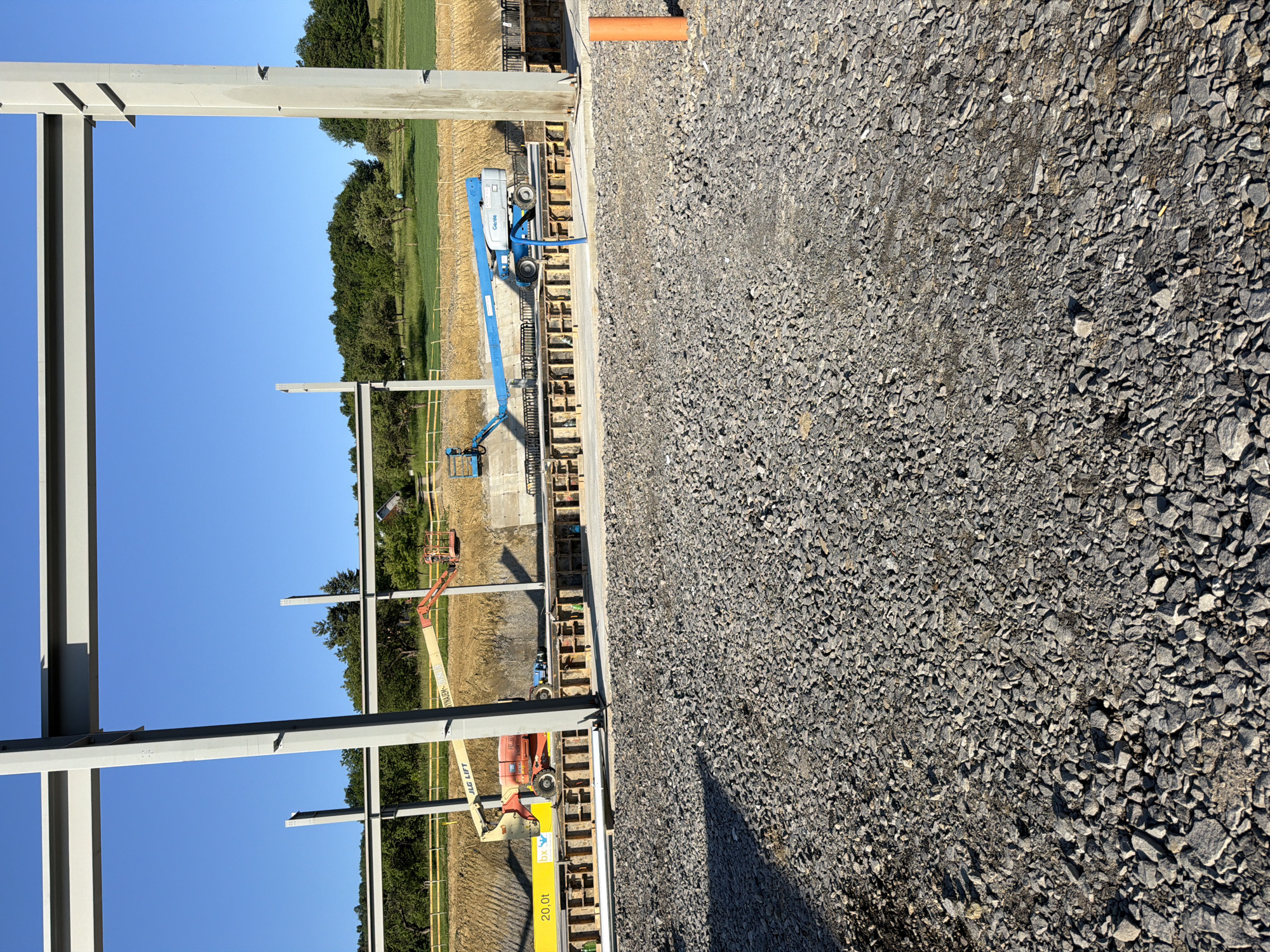 An outdoor, low-angle, vertical shot captures a construction site under a clear blue sky. The foreground is filled with a bed of grey gravel. In the middle ground, a large, unfinished steel building frame dominates the scene, with a grey I-beam column on the right and the edge of another on the left. Beyond the frame, there is a dirt embankment, a yellow safety fence, and a green field dotted with trees. A construction crew is present, operating two aerial work platforms: a red JLG Lift on the left and a blue Genie lift on the right. The red lift is positioned near a yellow sign that reads "20,0t bx". The blue lift is parked near a concrete structure. In the far background, a line of trees separates the construction site from a residential area with a house visible. The sun casts long shadows across the gravel in the foreground. The image is located in Stróża, Poland.