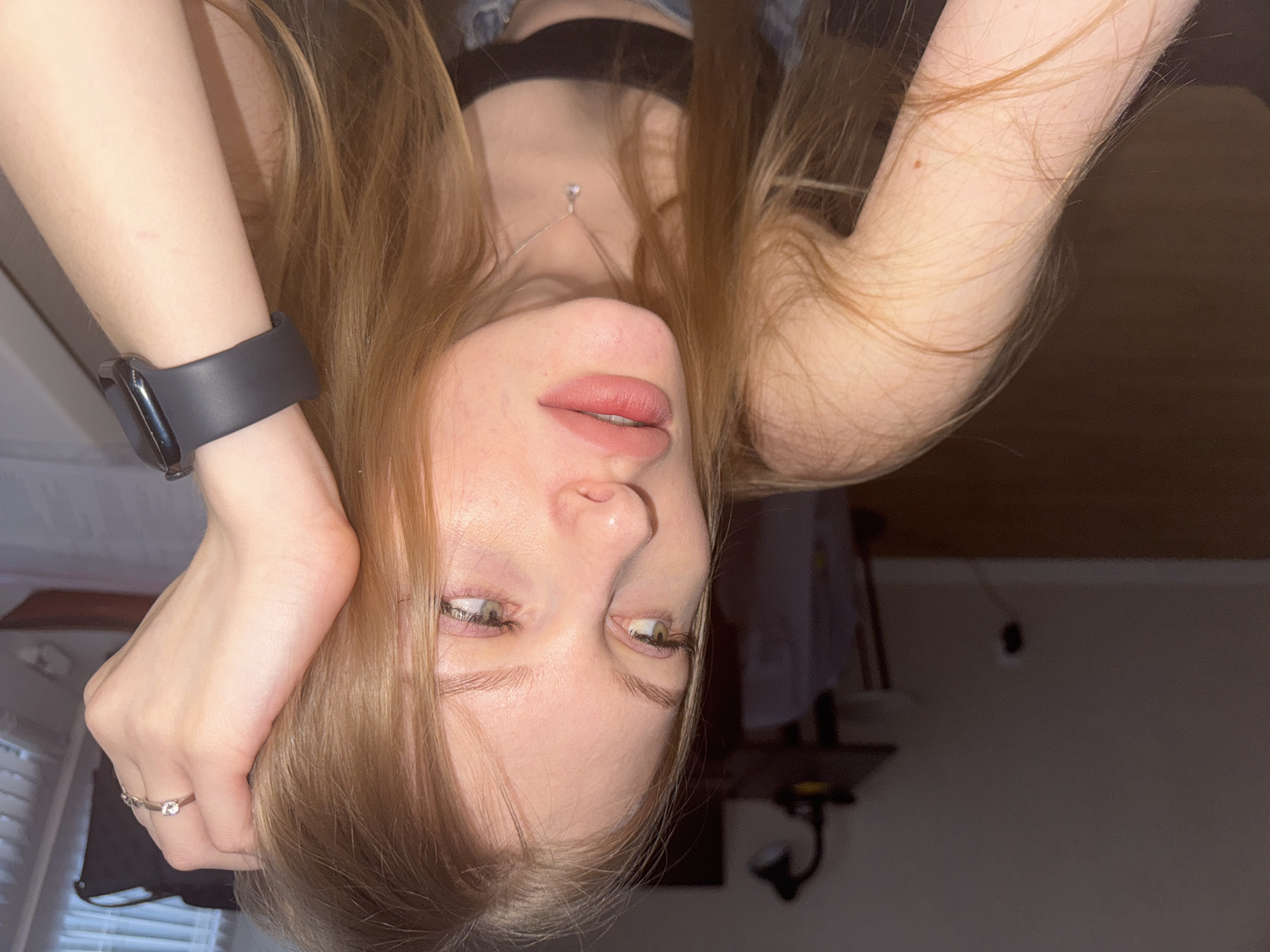 The image is an upside-down, close-up portrait of a young woman with long, light brown hair. Her head is positioned at the bottom of the frame, with her hair flowing upwards. She has fair skin, full lips with light pink lipstick that are slightly parted, and green or hazel eyes looking towards the top of the frame. Her nose is prominent, and her eyelashes are visible.
She wears a dark smartwatch with a black band on her left wrist, and a faint scar is visible on her forearm above the watch. On the ring finger of her left hand, she has a silver-colored ring featuring a clear, round stone. This hand is raised, possibly resting near her head or holding some hair. Around her neck, a delicate silver-toned necklace with a small, clear, teardrop-shaped pendant is visible. She appears to be wearing a dark top, possibly a bra or crop top. A small, dark mole or freckle is present on her right forearm.
The setting is indoors, characterized by soft, dim lighting, suggesting evening or a room with limited natural light. The background is blurred, showing hints of blinds or a window on the left side and indistinct furniture or objects, possibly a lamp or hanger, against a plain wall on the right. There is no visible text in the image. The overall impression is a posed, artistic self-portrait. This indoor scene is potentially set in Perm, Russia.