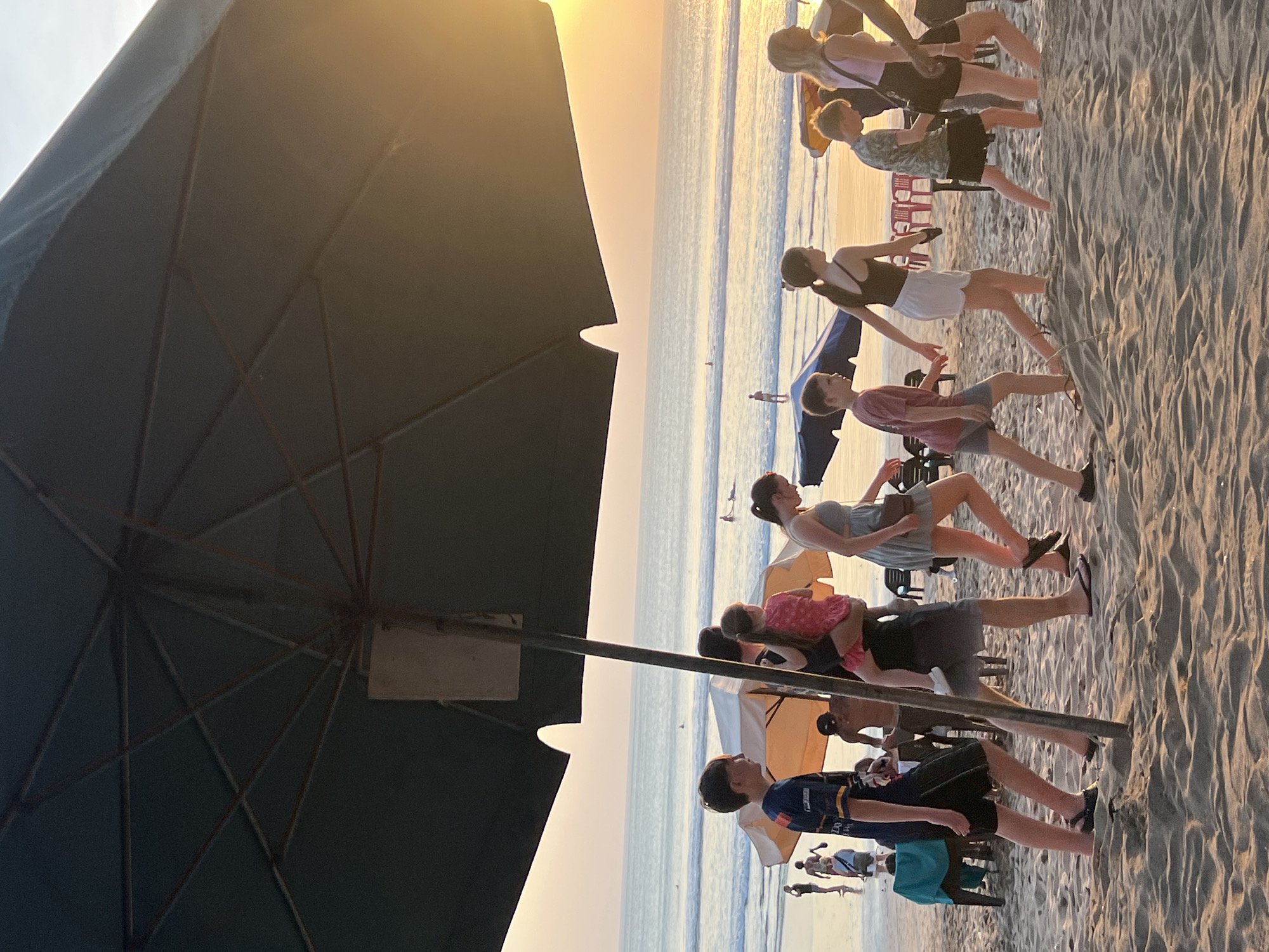 A group of people walk along a sandy beach in Kuta, Indonesia, under a large, dark umbrella. The sun is setting over the ocean, casting a warm glow and reflecting on the water. Several individuals, including children and adults, are moving from left to right across the frame. Some are wearing casual beach attire. In the background, there are beach umbrellas, chairs, and people in the ocean, suggesting a typical evening scene at this popular tourist destination. The sand shows ripples from the wind or footprints.