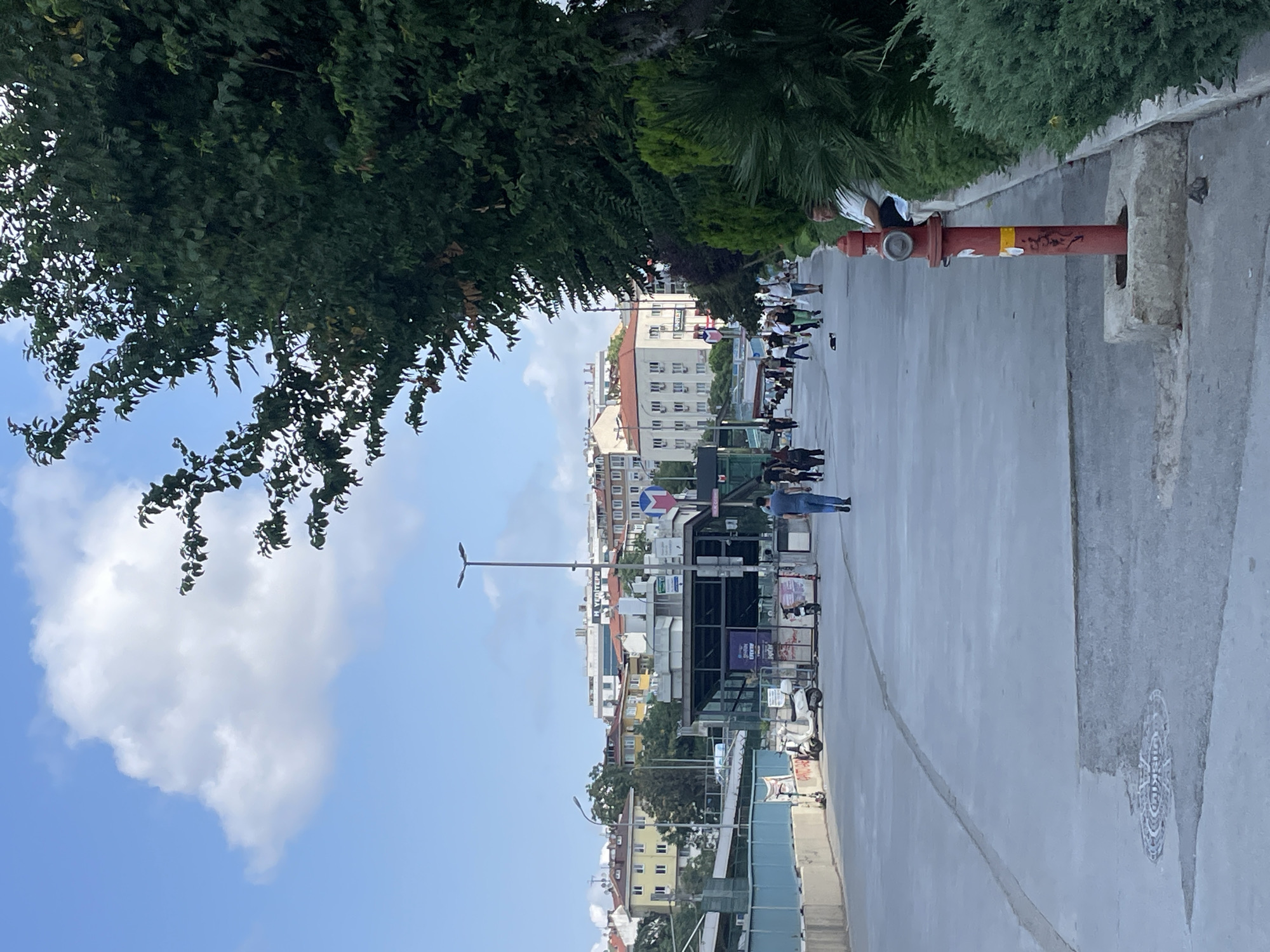 The image captures a street scene in Kadıköy, Turkey, on a clear day with scattered clouds. In the foreground, a wide, paved walkway runs horizontally across the frame, with a man in blue jeans and a dark t-shirt walking away from the camera towards the center of the image. To the right, a bright red fire hydrant stands on a concrete base, with a man in a white t-shirt and jeans seated on a low wall behind it, facing away from the camera. Lush green bushes and trees line the right side of the walkway.
In the middle ground, the walkway opens up to an area with several parked scooters and a small kiosk or building with signage. A group of people are walking in the same direction as the primary subject, with some standing and conversing. Beyond this area, the cityscape of Kadıköy unfolds, featuring multi-story buildings with a mix of architectural styles, including some with yellow facades and others with white and cream colors. The sky above is a bright blue, with a large, fluffy white cloud dominating the upper left portion of the image. The scene suggests a public space in an urban environment, likely a promenade or pedestrian area.
