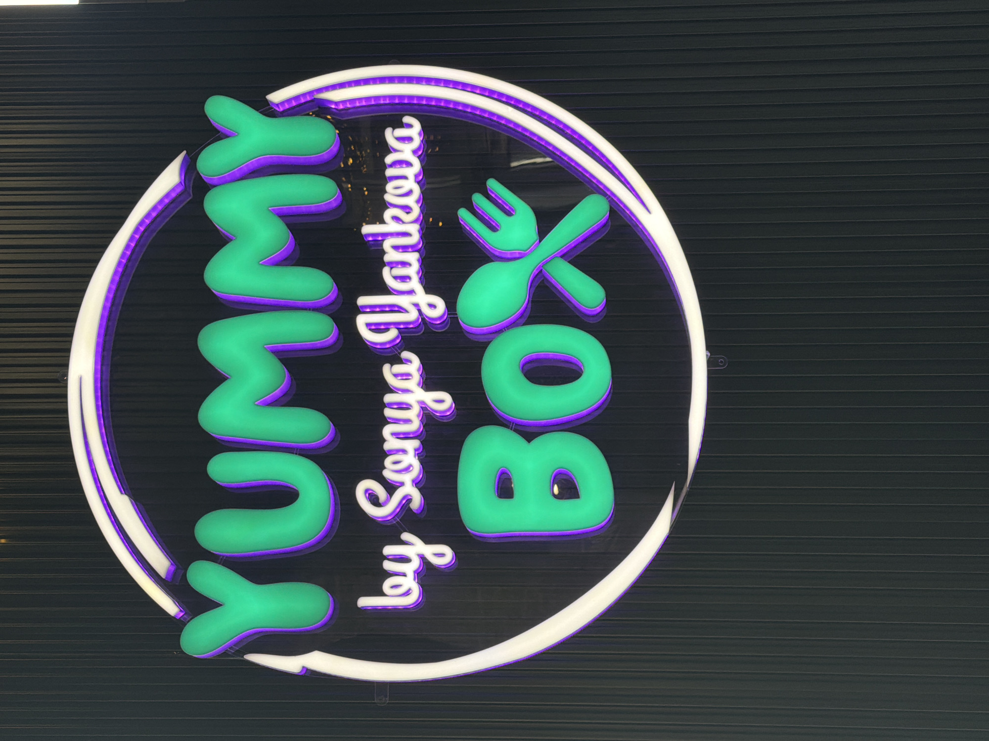 A lit sign for "Yummy Box by Sonya Yankova" is mounted on a dark, horizontal-striped surface. The sign features green, cartoon-like letters for "Yummy Box" with a purple outline. A stylized, green spoon and fork are crossed over the word "Box". The words "by Sonya Yankova" are in a white, script-like font. The entire sign is framed by a white circle that has a purple accent glow. The image appears to be taken indoors. Sofia, Bulgaria is mentioned as the location.
