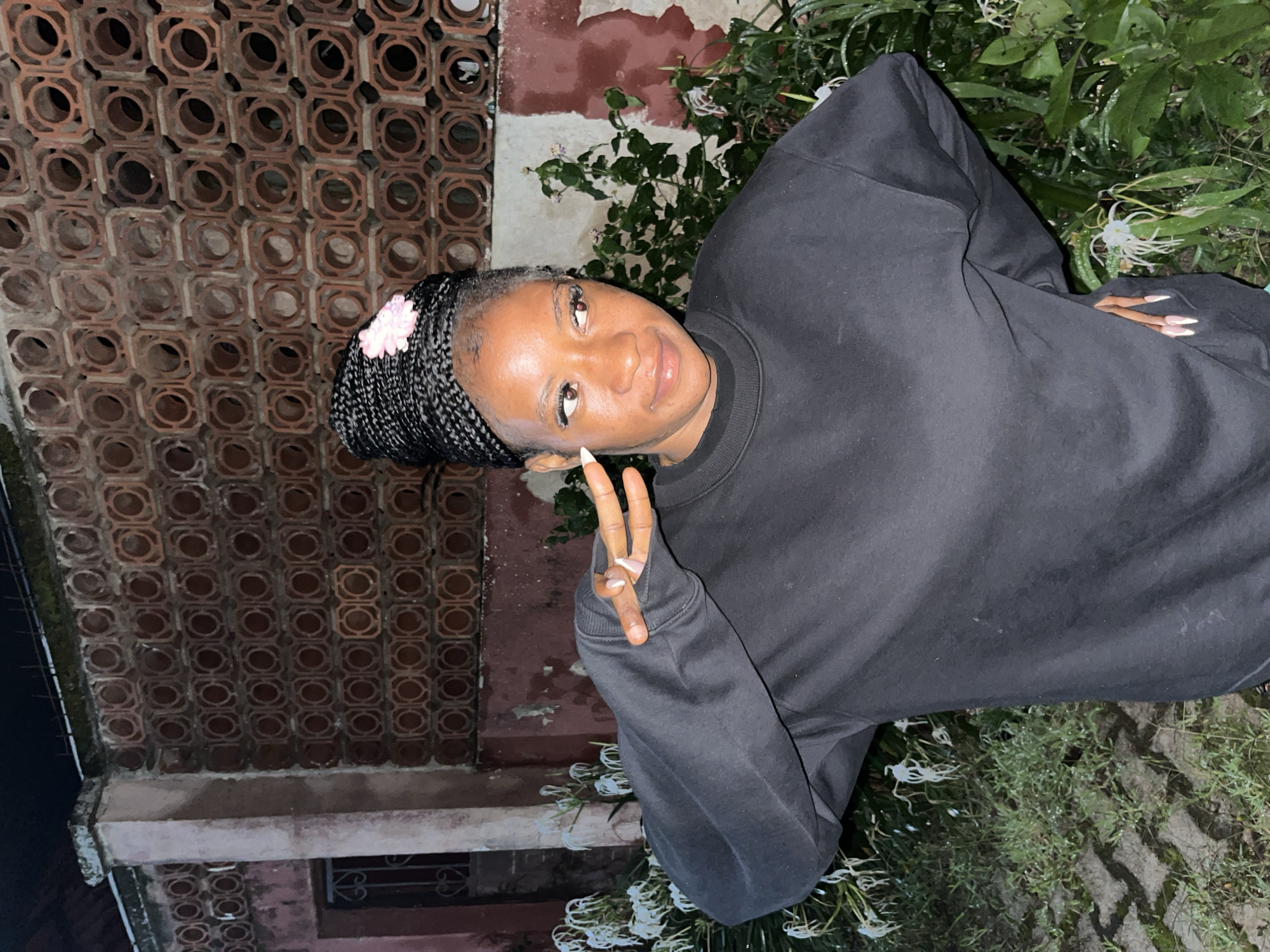 A young woman with braided hair, adorned with a small pink flower, stands in an outdoor setting at night. She is wearing a black sweatshirt and has her right hand raised, forming a peace sign with her fingers. Her gaze is directed to her right, and she has a subtle smile. Behind her, a textured brick wall with circular openings serves as a backdrop, partially obscured by lush green foliage and white flowers. The ground appears to be paved with interlocking stones. The overall lighting suggests it is nighttime, possibly in a residential area or courtyard within Jos, Nigeria.