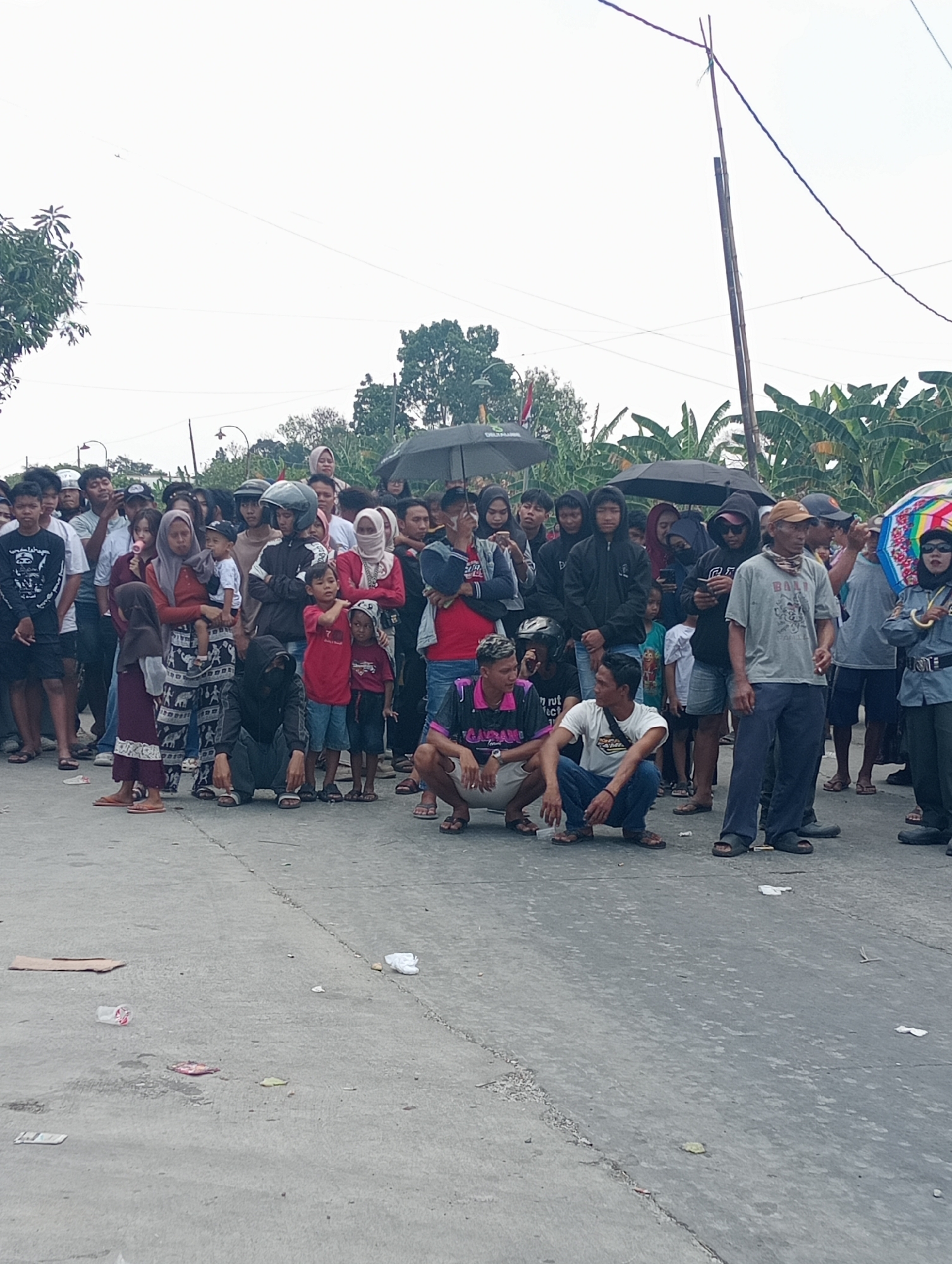 A crowded street scene unfolds in Doreng, Indonesia, under an overcast sky. A diverse group of individuals, including men, women, and children, are gathered, some standing and others squatting on a paved road. Many are looking in the same direction, suggesting a shared point of focus or event. Several people are holding umbrellas, indicating potential rain or strong sun.
The environment appears to be a residential or semi-urban area, with banana trees visible in the background. The road is worn and shows signs of debris, including discarded paper and plastic. A bamboo pole with electrical wires stretches across the frame, a common sight in many Indonesian locales. The overall atmosphere is one of community gathering, with a sense of anticipation or observation. The clothing worn by the individuals is casual, reflecting the tropical climate. The presence of multiple umbrellas suggests a communal response to weather conditions. The scene is captured from a slightly elevated perspective, looking down onto the assembled crowd and the street.