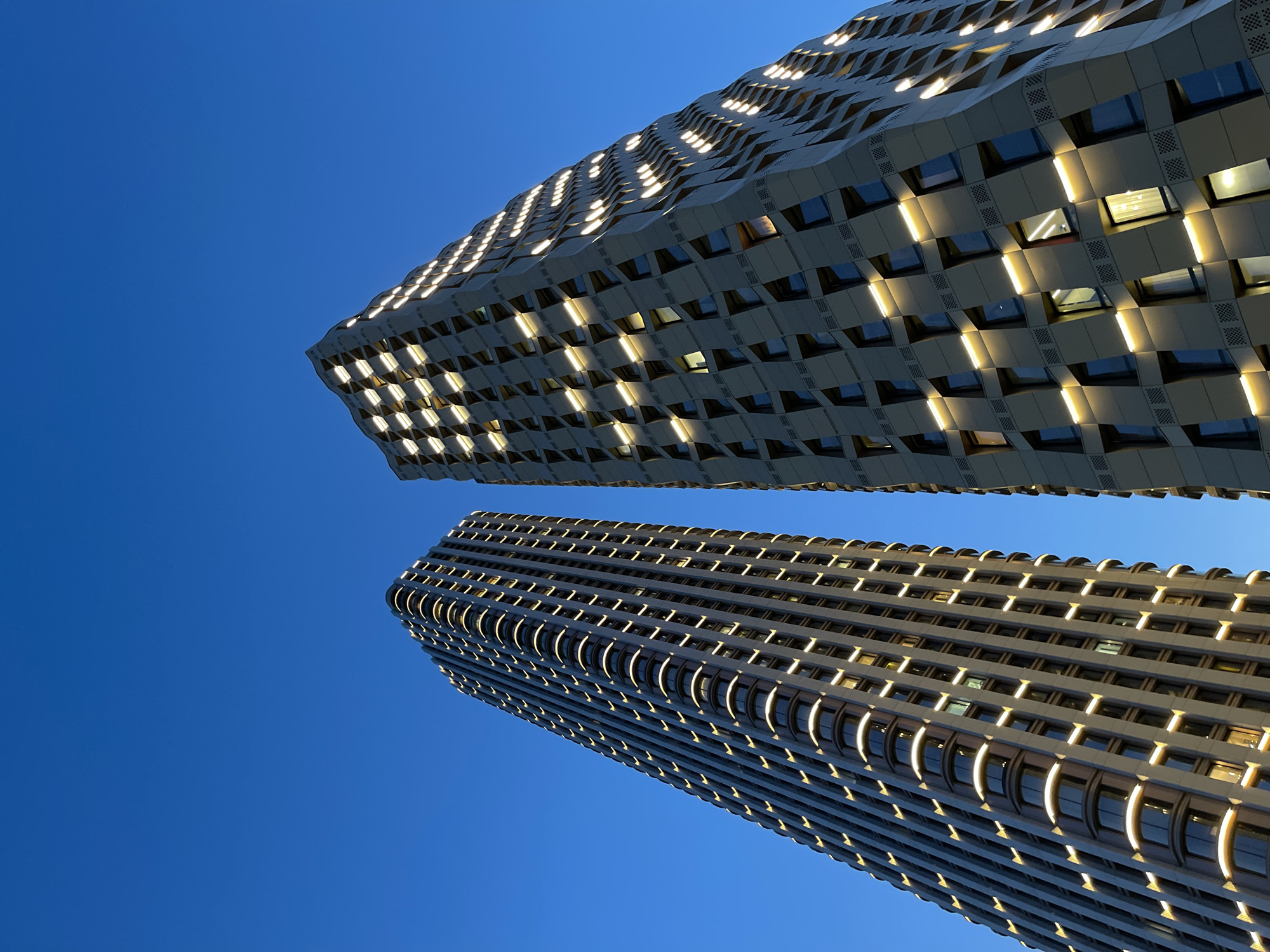 A low-angle, upward-facing shot captures two tall buildings against a clear, deep blue sky. The buildings are modern with a geometric facade, and illuminated with rows of windows that are lit from the inside, giving a grid-like pattern of light and shadow. The building in the background has a pattern of angular segments, while the building closer to the viewer has curved window frames. The lighting emphasizes the textures of the buildings. The location is indicated as Khimki, Russia, but the scene contains no indicators, signs, or people.