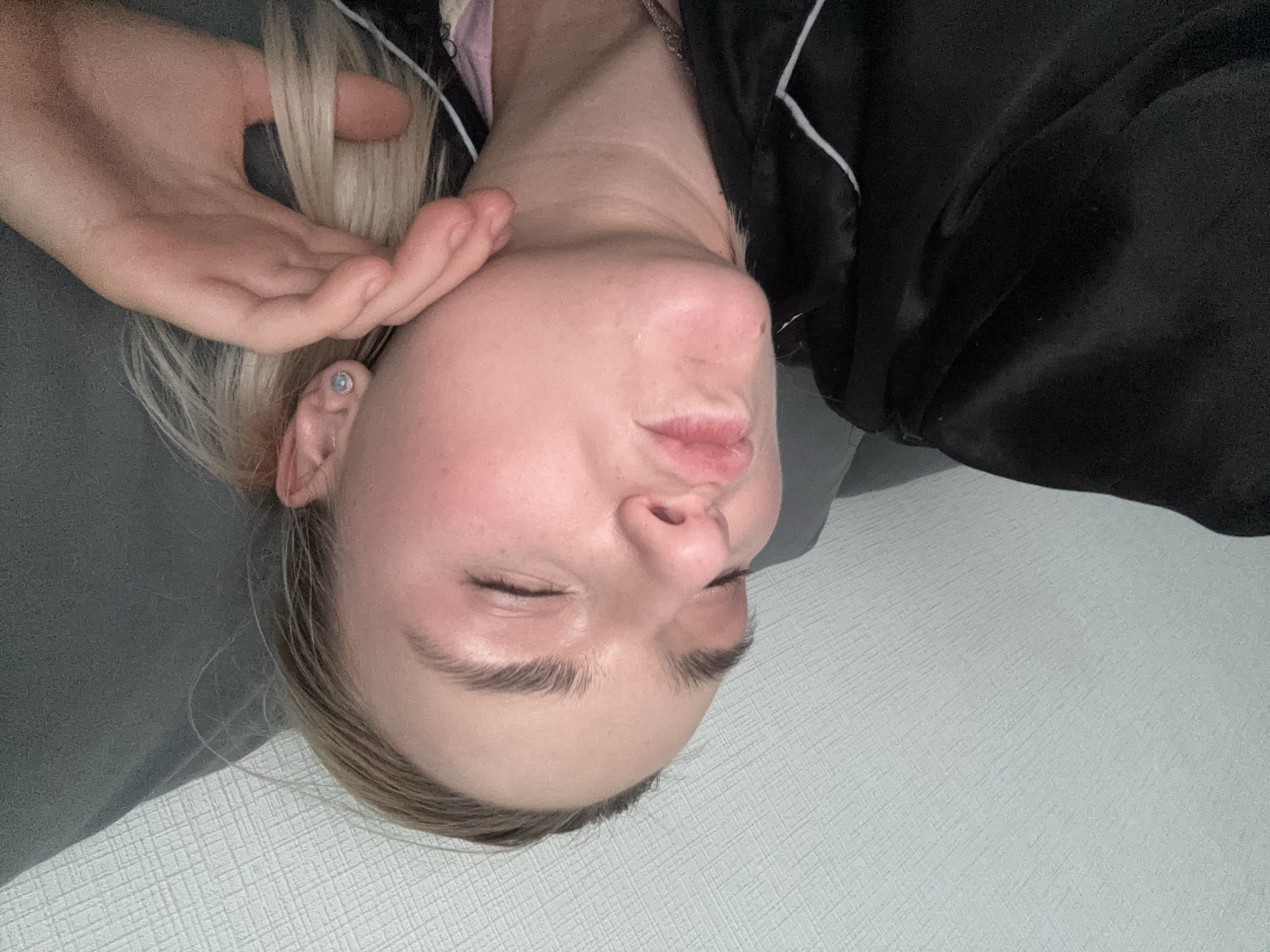 A woman with blonde hair is lying on a light gray surface, possibly a bed or sofa. She is wearing a black garment with white trim. Her eyes are closed and she is holding her hand near her face. Her facial expression is relaxed, and she may be asleep or resting. The location is unknown, Russia.