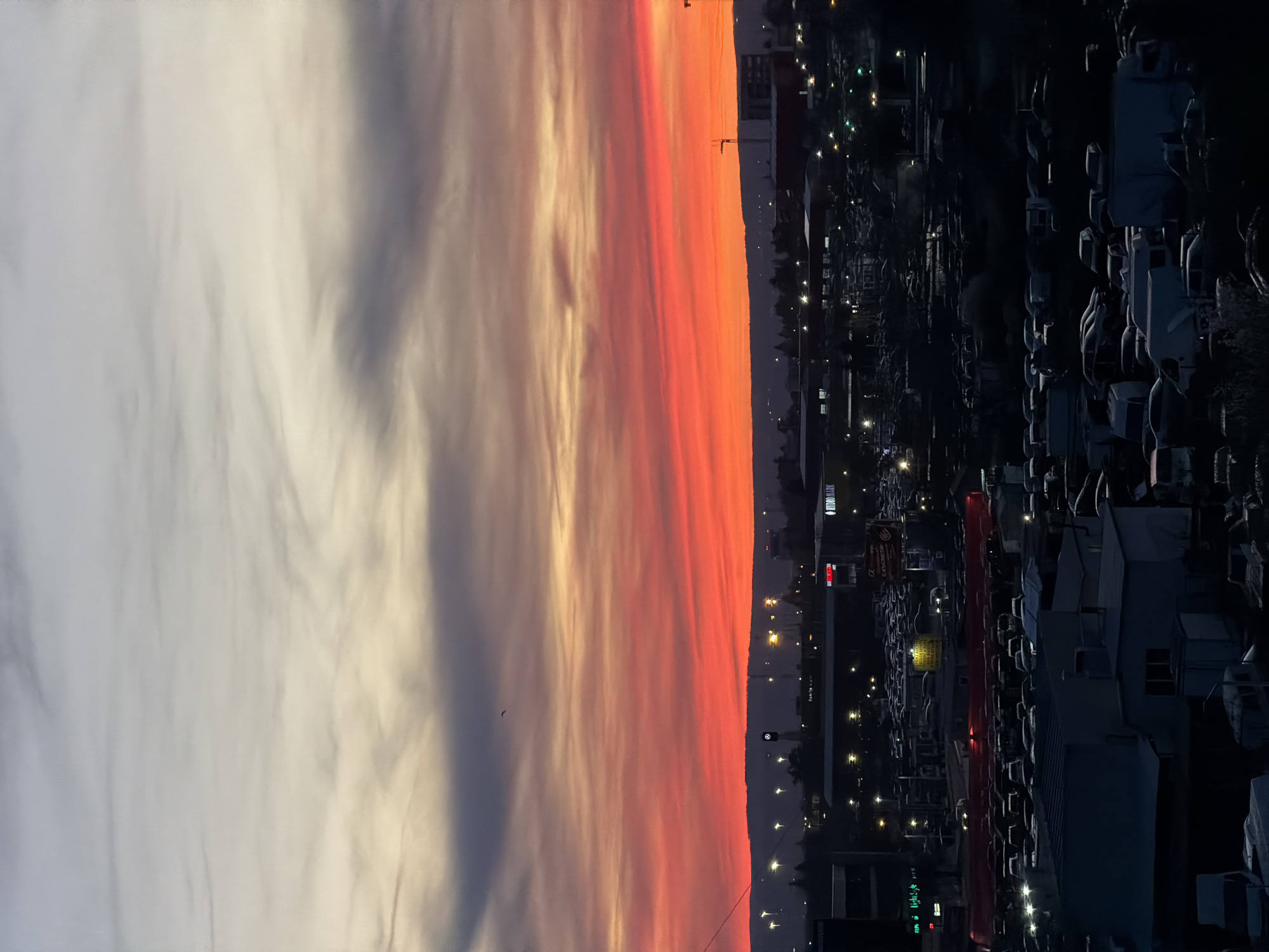 The image depicts a vibrant sunset over an urban area, likely taken from an elevated position. The sky is divided into two distinct parts: the left side is dominated by white and grey clouds, while the right side showcases a dramatic display of orange and red hues. Below the horizon, a cityscape unfolds, characterized by numerous buildings, streetlights, and vehicles. The scene appears to be captured during dusk or twilight, as evidenced by the combination of fading daylight and artificial lighting.
Based on visual cues, it is impossible to ascertain the exact location of the image in Varna, Bulgaria.