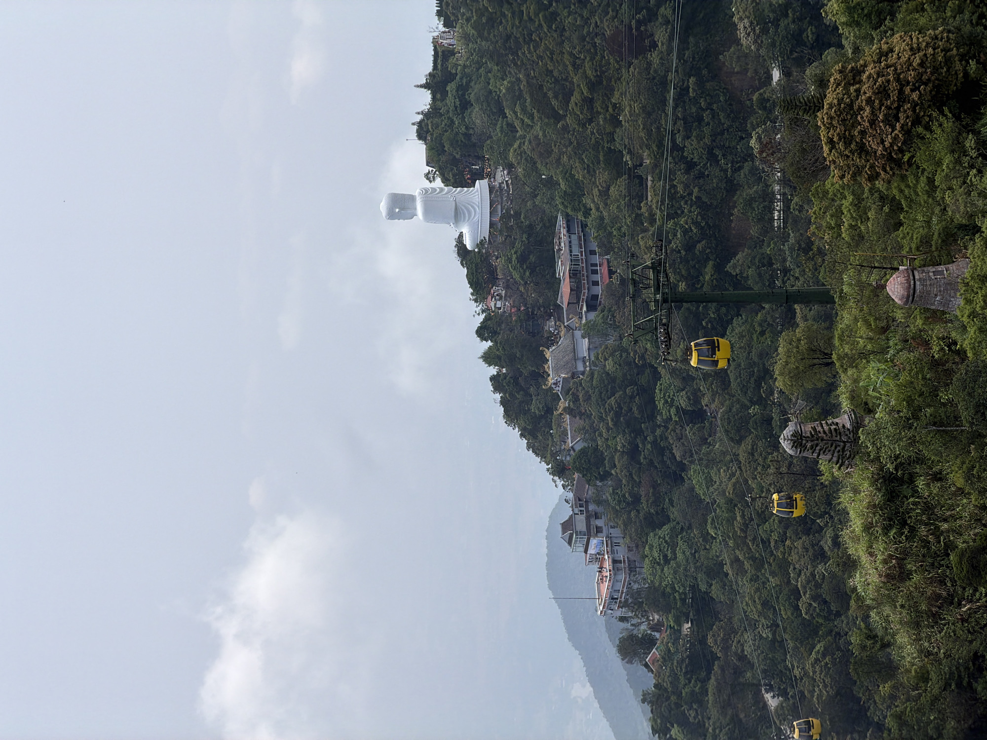 The image depicts a scenic view of Ba Na Hills in Đà Nẵng, Vietnam. A large white Buddha statue is visible atop the mountain, partially obscured by mist. A cable car system with yellow gondolas traverses the landscape, with several cars in motion. The hillside is densely covered with lush green trees and vegetation. Buildings and structures are dotted along the slope. The sky is overcast with white clouds.