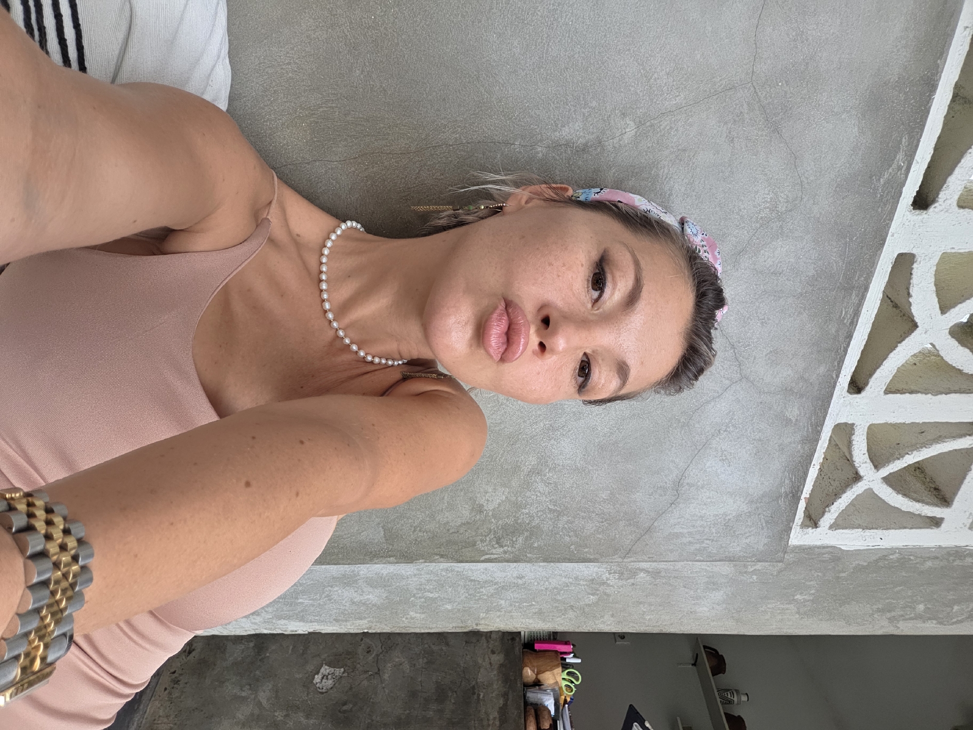 A woman with blonde hair tied back with a floral headband and wearing a pearl necklace and a pink dress is leaning against a gray concrete wall. She is puckering her lips as if for a kiss and her left arm is bent, resting on her shoulder. A gold and silver bracelet adorns her wrist. Behind her, a white decorative screen is visible, and a shelf with various items can be seen on the right side of the image. The setting appears to be outdoors, possibly in a cafe or restaurant in North Kuta, Indonesia.