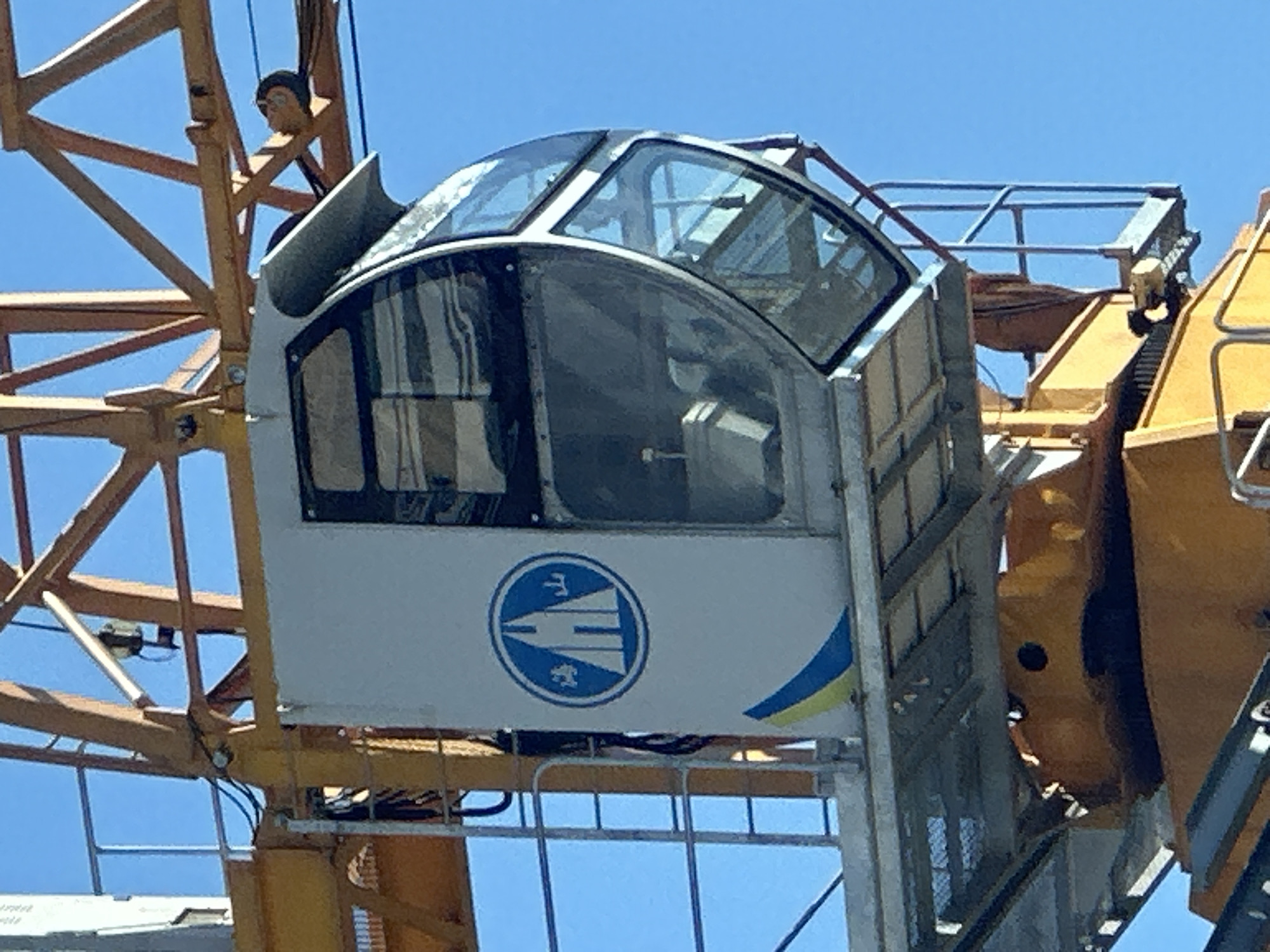 The image shows a close-up of a construction crane's operator cabin against a clear blue sky. The cabin is white with a curved glass front and a blue and yellow stripe design on its side. A blue circular logo with a white building and Chinese characters is visible on the cabin's exterior. The crane itself is yellow and appears to be in an active construction site. There are no people clearly visible within the cabin. The image does not provide specific details about the location in Kyiv, Ukraine, beyond it being a construction site.