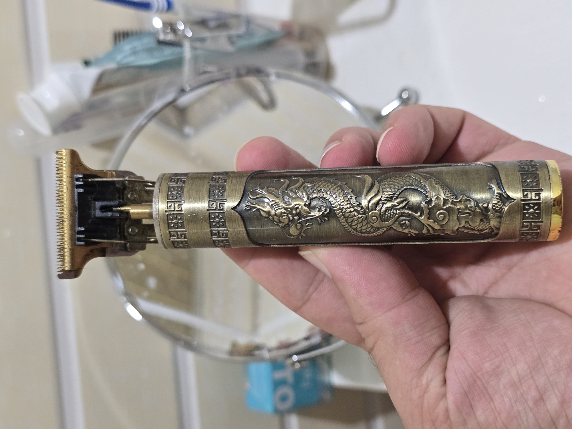 A hand holds a close-up of a vintage-style metal hair clipper with intricate dragon detailing. The clipper is bronze-colored and appears to be an electric trimmer with a sharp metallic blade at the top. The background is blurred, showing a bathroom sink area with a mirror, a toothbrush holder containing toiletries like toothpaste and toothbrushes, and a small blue box with white lettering. The overall scene suggests a personal grooming item in a domestic setting. The city of Qaratau, Kazakhstan is not visible or implied in the image.