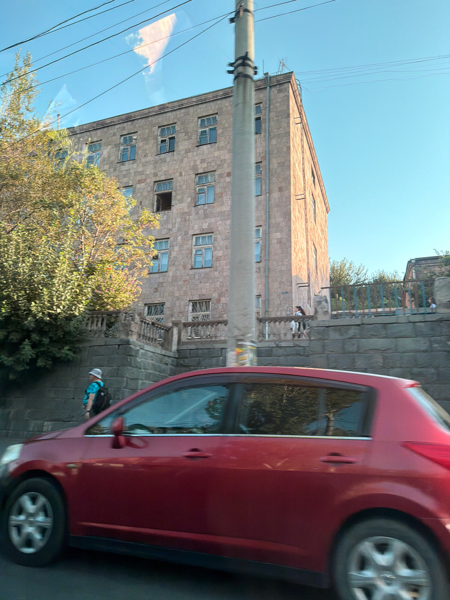 The image shows a street scene in Yerevan, Armenia, featuring a red car in motion in the foreground. Behind the car, a multi-story building constructed from light-colored stone blocks is visible. The building has numerous windows, some open and some closed. Adjacent to the building is a set of stone stairs with decorative railings. A person wearing a light-colored hat, a blue patterned shirt, and a backpack is walking up the stairs. A tall concrete utility pole stands to the left of the building. Trees with yellowing foliage are also present. The sky is clear and blue, with some reflections visible on the windows of the car.