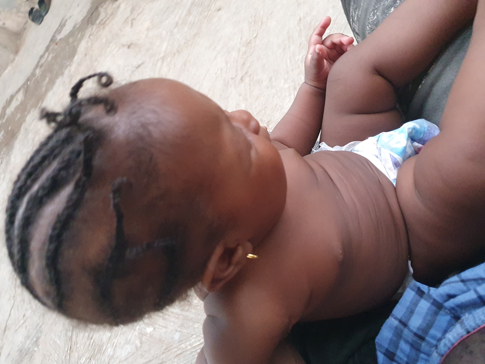 An infant with a braided hairstyle and a gold earring is lying on their back. The baby is wearing a diaper. The location is assumed to be Lokoja, Nigeria. Other individuals are present but only their clothing is partially visible. The baby is laying on some type of material, and the background shows a textured concrete surface.