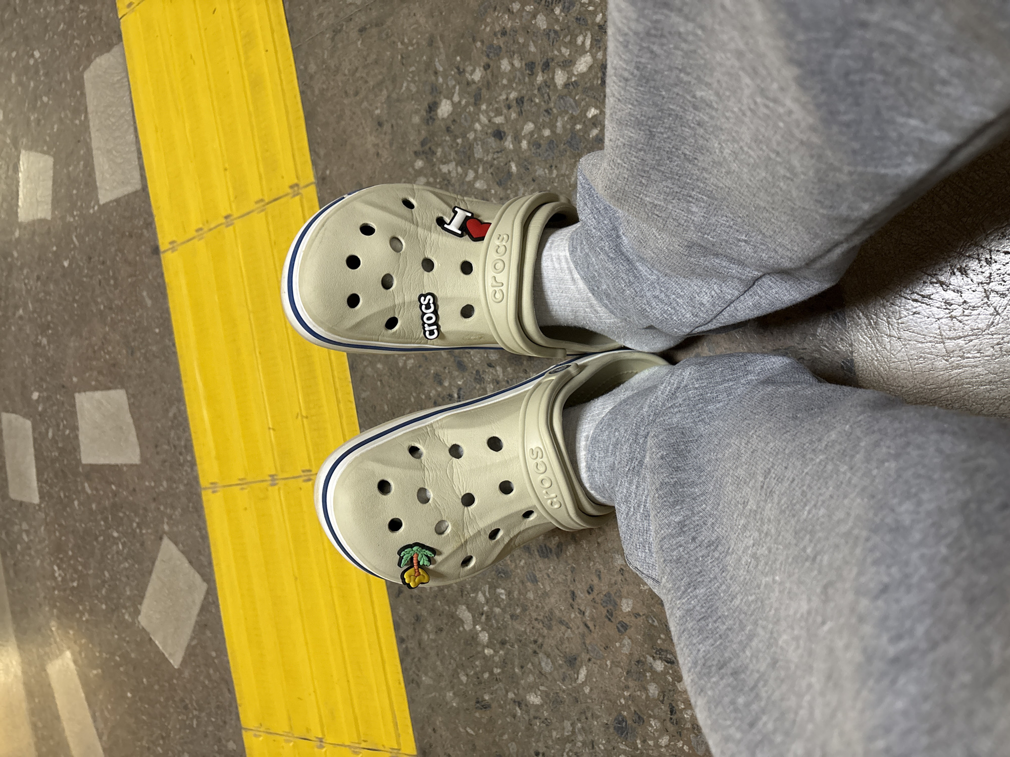 The image shows a pair of light beige Crocs with blue trim, adorned with two Jibbitz charms: one is an "I heart" symbol and the other is a palm tree with a yellow fruit. The Crocs are worn with light grey sweatpants and white socks. The scene is set on a speckled grey floor with a prominent, raised yellow line, possibly a tactile warning strip. The lighting suggests it is daytime, and the setting appears to be an indoor or covered area due to the smooth, manufactured flooring and the absence of natural outdoor elements. The "Crocs" logo is visible on both shoes.