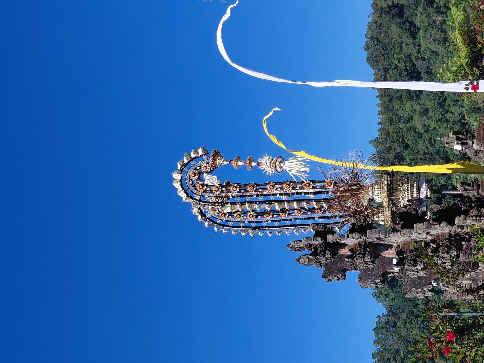 The image depicts a vibrant scene with a clear blue sky dominating the upper portion. In the foreground, a tall, ornate, tiered structure, possibly a temple decoration or a ceremonial arch, is adorned with intricate carvings and colorful elements. To the left of this structure, a part of a weathered, stone building with multiple levels and decorative carvings is visible, suggesting an ancient or traditional architectural style.
Flanking the central decoration are two large, flowing banners. A bright yellow banner curves gracefully to the left, while a stark white banner drapes to the right. In the background, a dense forest of lush green trees covers a hillside. The overall impression is one of cultural significance and natural beauty, possibly captured during a festival or a special occasion in a location like Baturiti, Indonesia, known for its temples and natural landscapes. Red flowers are also visible in the lower left corner of the image.