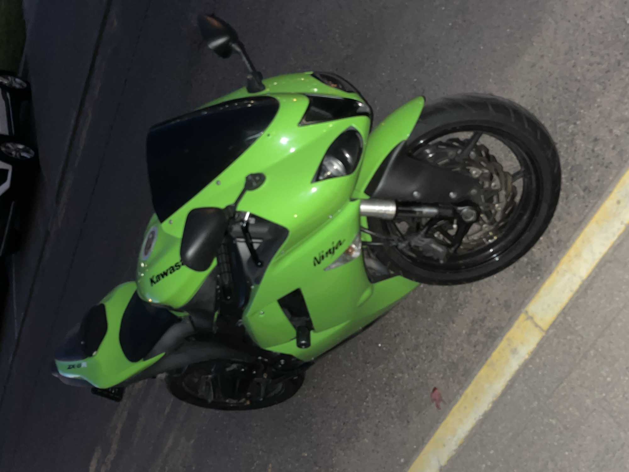 An overhead shot captures a bright green Kawasaki Ninja ZX-6R sport motorcycle parked on an asphalt surface, possibly at dusk or night given the muted lighting. The motorcycle is the central focus, angled diagonally from the upper left to the lower right of the frame, and appears to be in pristine condition.
The bike features prominent green fairings with black accents, including the windscreen, seat, and handlebars. Visible text on the fairings clearly reads "Kawasaki" and "Ninja," with "ZX-6R" also discernable on the lower front fairing. Black multi-spoke wheels, brake discs, and a front suspension system are visible. A black frame slider is present on the lower side.
To the left of the motorcycle, part of a dark car is visible, suggesting a parking lot or street setting. The ground is dark gray asphalt, and a distinct yellow painted curb or line runs along the right side of the image, separating the pavement from an unseen area. The overall impression is a vibrant motorcycle at rest in an urban or suburban outdoor environment in Minsk, Belarus.