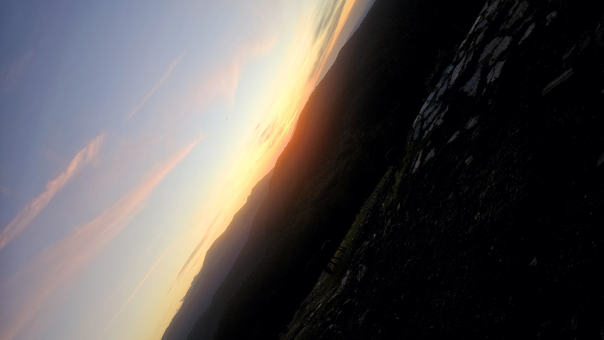 The image depicts a landscape scene, likely at dawn or dusk, characterized by a diagonal orientation. The sky transitions from a pale blue at the top to a warm yellow and orange near the horizon, with delicate pink clouds streaking across. Silhouetted against the bright sky are the layered shapes of distant mountains, their forms softened by atmospheric haze. In the foreground, the dark mass of a hillside slopes downwards, appearing rocky and uneven. A faint outline of what could be a horse or similar animal is visible grazing on a patch of green grass on the hillside. The overall impression is one of natural beauty and tranquility, with the low light casting deep shadows. The location in Simeonovets, Bulgaria, is not explicitly identifiable within the visual elements of the image itself.