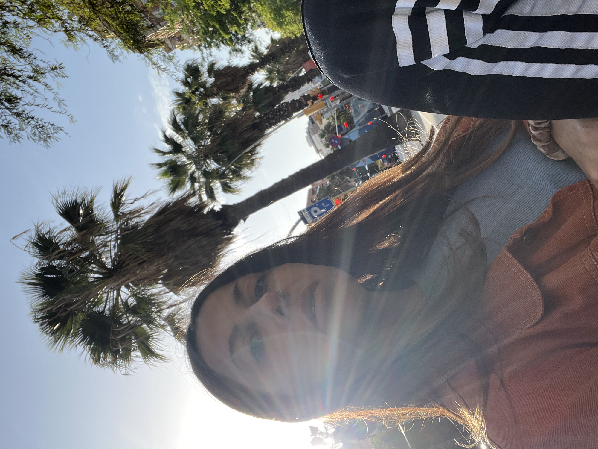 The image captures a bright, sunny outdoor scene, likely taken as an upward-facing selfie or from a low angle. The primary subject is a person, appearing to be a young woman, with long dark brown hair, looking directly towards the camera with a neutral to slightly smiling expression. She has a subtle nose piercing. The strong sunlight creates a prominent lens flare across the right side of her face and hair.
She is dressed in a light blue top, layered with a reddish-brown jacket. Part of a black garment with distinctive white parallel stripes, resembling Adidas sportswear, is visible in the upper right background. A light-colored scrunchie can be seen on her wrist or arm.
The setting is an urban environment, characterized by tall palm trees that line what appears to be a street or boulevard, with their fronds silhouetted against a clear, light blue sky. In the background, beyond the palm trees, parts of buildings and traffic are visible, including red traffic lights on cars and a partially obscured blue street sign with a white 'P' symbol, indicating parking. Other green foliage from various trees frames the upper parts of the sky.
The strong sunlight suggests it is midday or early afternoon, with clear weather. The presence of palm trees and the general architecture align with the provided context of Barcelona, Spain. The image has a vertical perspective, suggesting an upward glance or a rotated capture, with the sky and tree canopy filling a significant portion of the frame.
