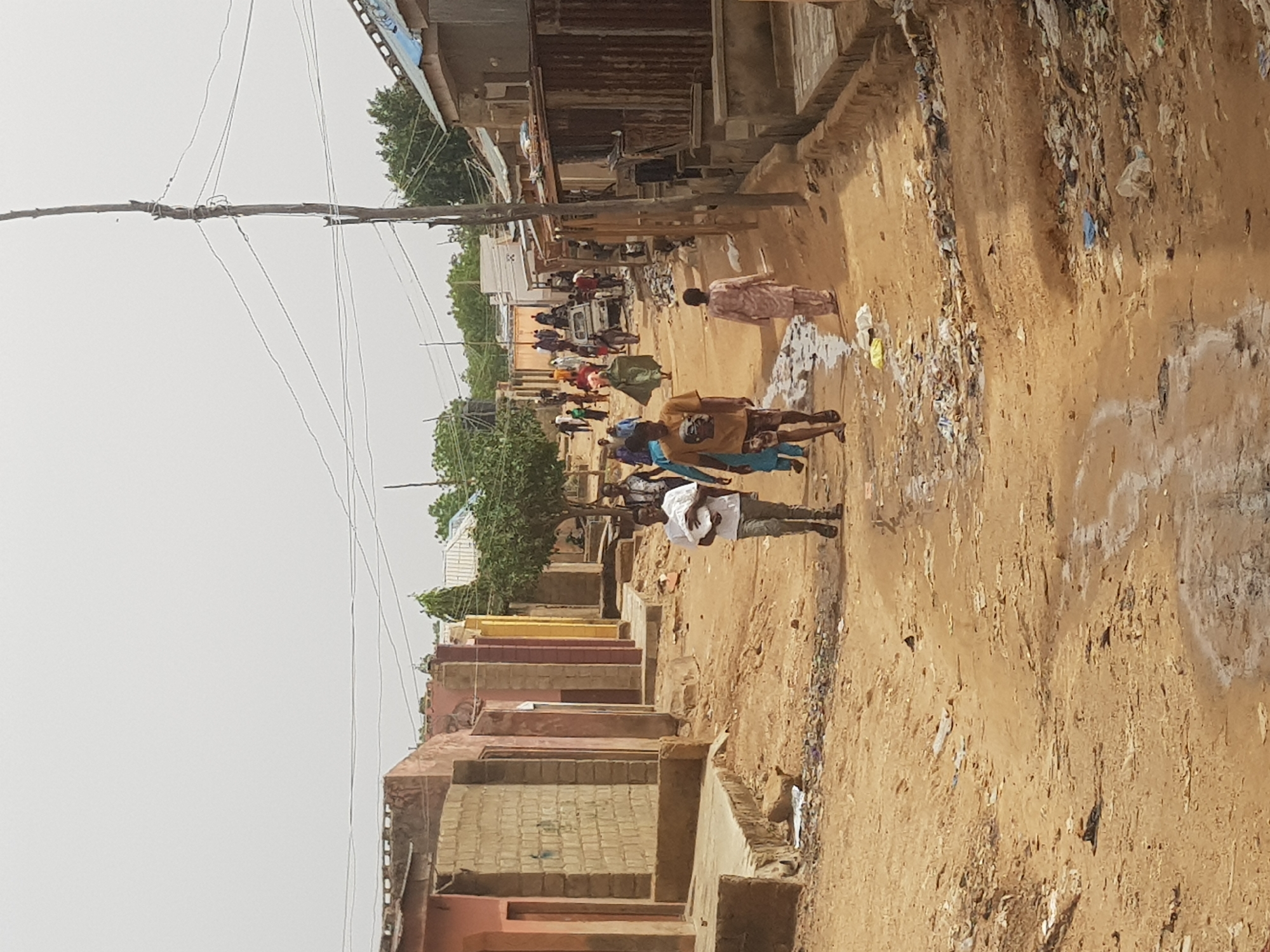 The image depicts a street in Dabawa, Nigeria. It is a dirt road with buildings on either side, some appearing to be unfinished or in a state of disrepair. There is a utility pole with visible wiring on the left. A group of people are walking along the road; their clothing is diverse. Litter and debris are scattered along the side of the road. A vehicle, potentially a small truck, is further down the road, surrounded by more people. The overall impression is one of an impoverished or underdeveloped area.