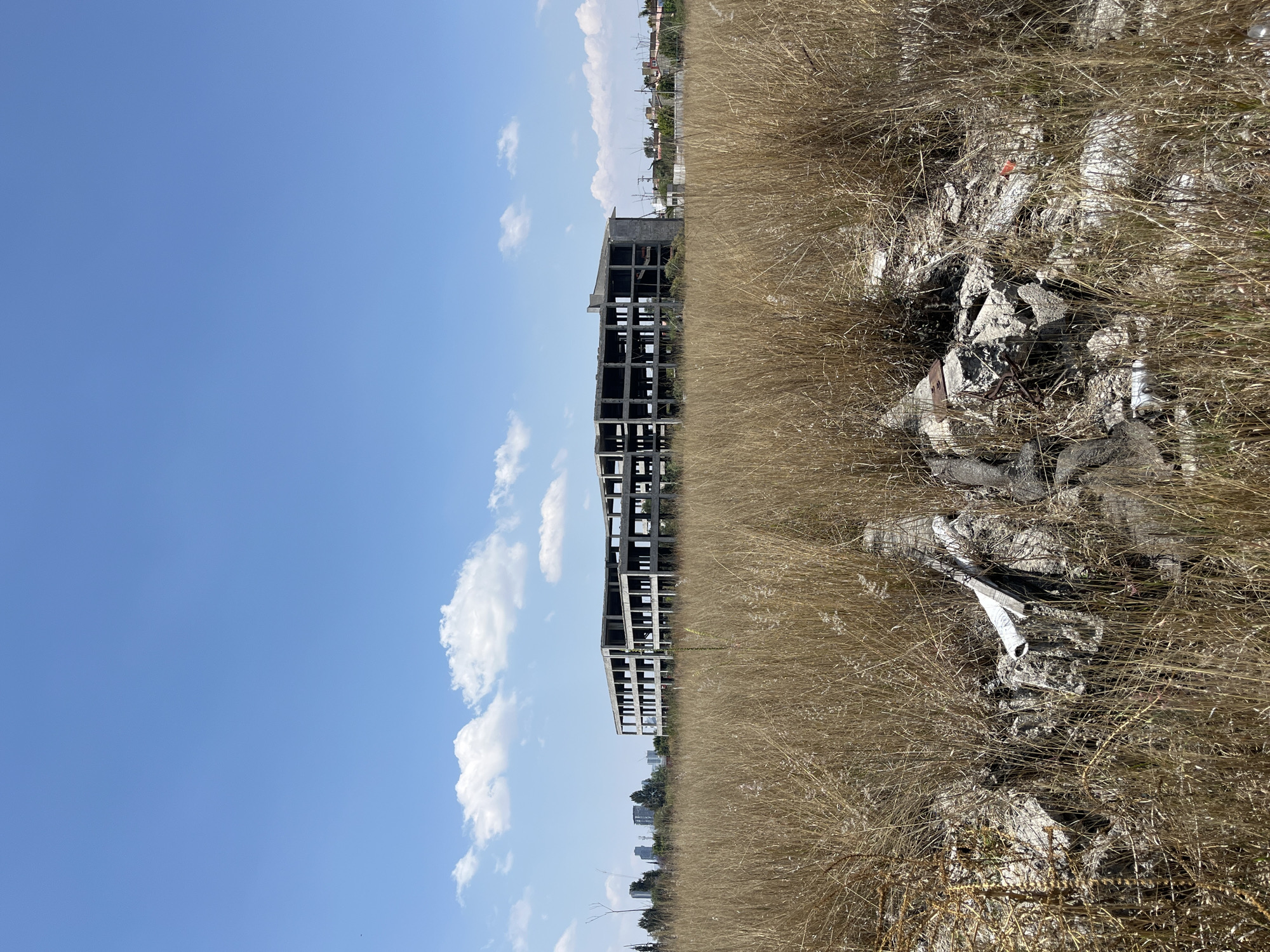 An exterior shot of an abandoned concrete building structure located in San Bernardino Tlaxcalancingo, Mexico. The sky is clear with scattered white clouds. To the right of the building is a field of dry brown grass, with debris and rubble visible.