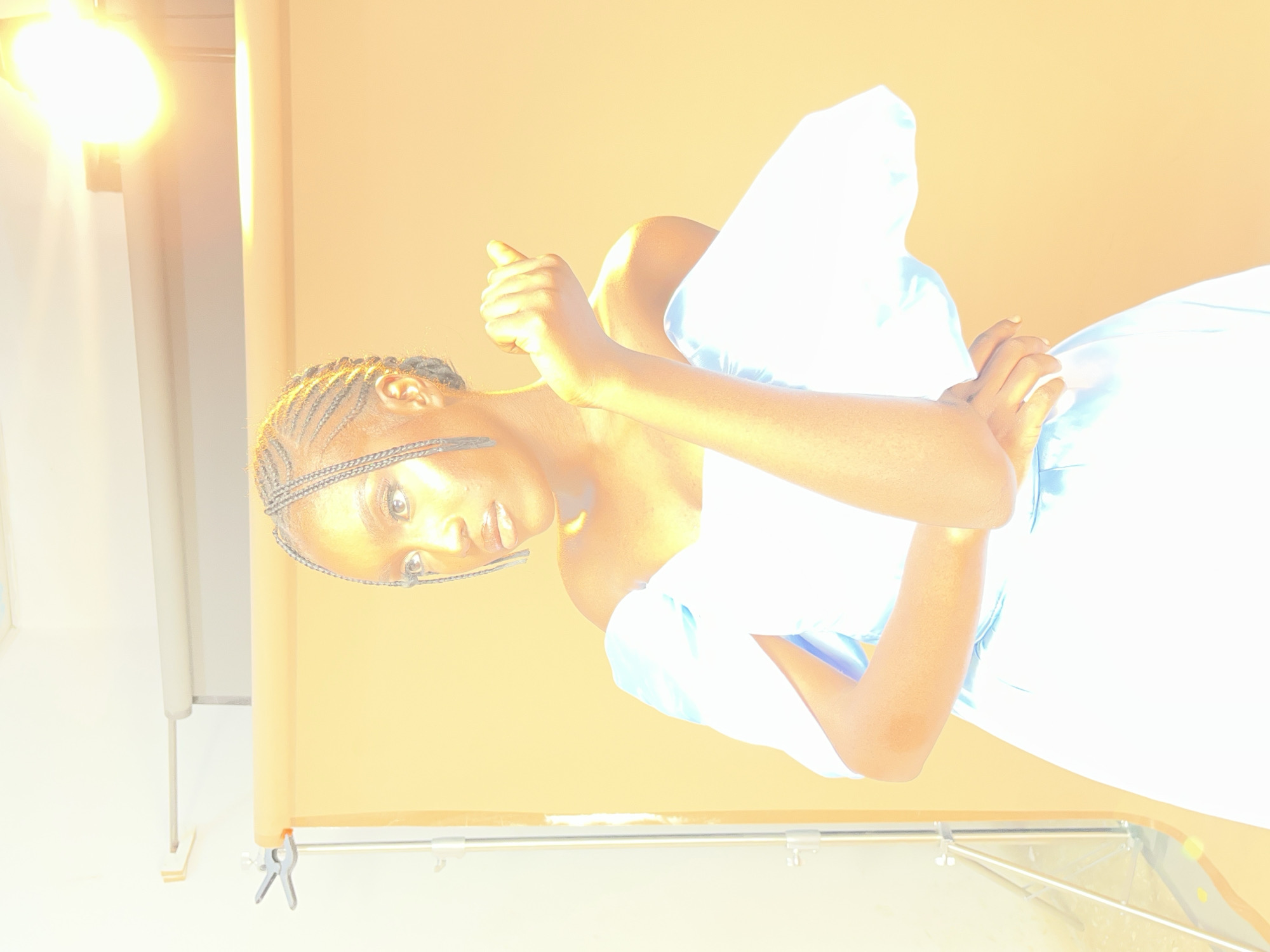 A young Black woman with braided hair, wearing a light blue off-the-shoulder dress, poses against a warm yellow backdrop. She is positioned in the foreground, slightly to the left of the frame, looking off to her left. Her right arm is bent, with her hand resting on her chest. A studio lighting rig is visible on the left side of the image, and a bright light source is positioned in the upper right corner. The scene appears to be a professional studio setup in Gura Top, Nigeria, indicated by the context provided.