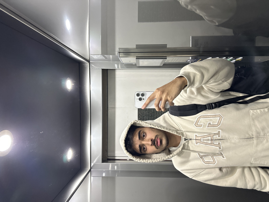 The image displays a single adult male taking a mirror selfie inside an enclosed space, likely an elevator. The male, with dark hair and a mustache, is shown from the upper torso, wearing a light beige zip-up hoodie featuring a cream-colored shearling-like lining in the hood. The word "GAP" is embroidered in large, outlined, light brown letters on the front of the hoodie. A dark-colored backpack is visible over his right shoulder. He holds a white smartphone with a multi-lens camera in his right hand, pointed towards the reflective surface to capture his image. The environment is characterized by silver-grey, highly reflective metallic or mirrored panels on the walls and ceiling. Two distinct circular recessed lights illuminate the ceiling. A portion of a silver-grey elevator door is visible on the left side, and a reflective panel with an indistinct control interface is present on the right. No identifiable architectural features, specific signage, or landmarks are visible within the image to confirm the precise location in Guimarães, Portugal.