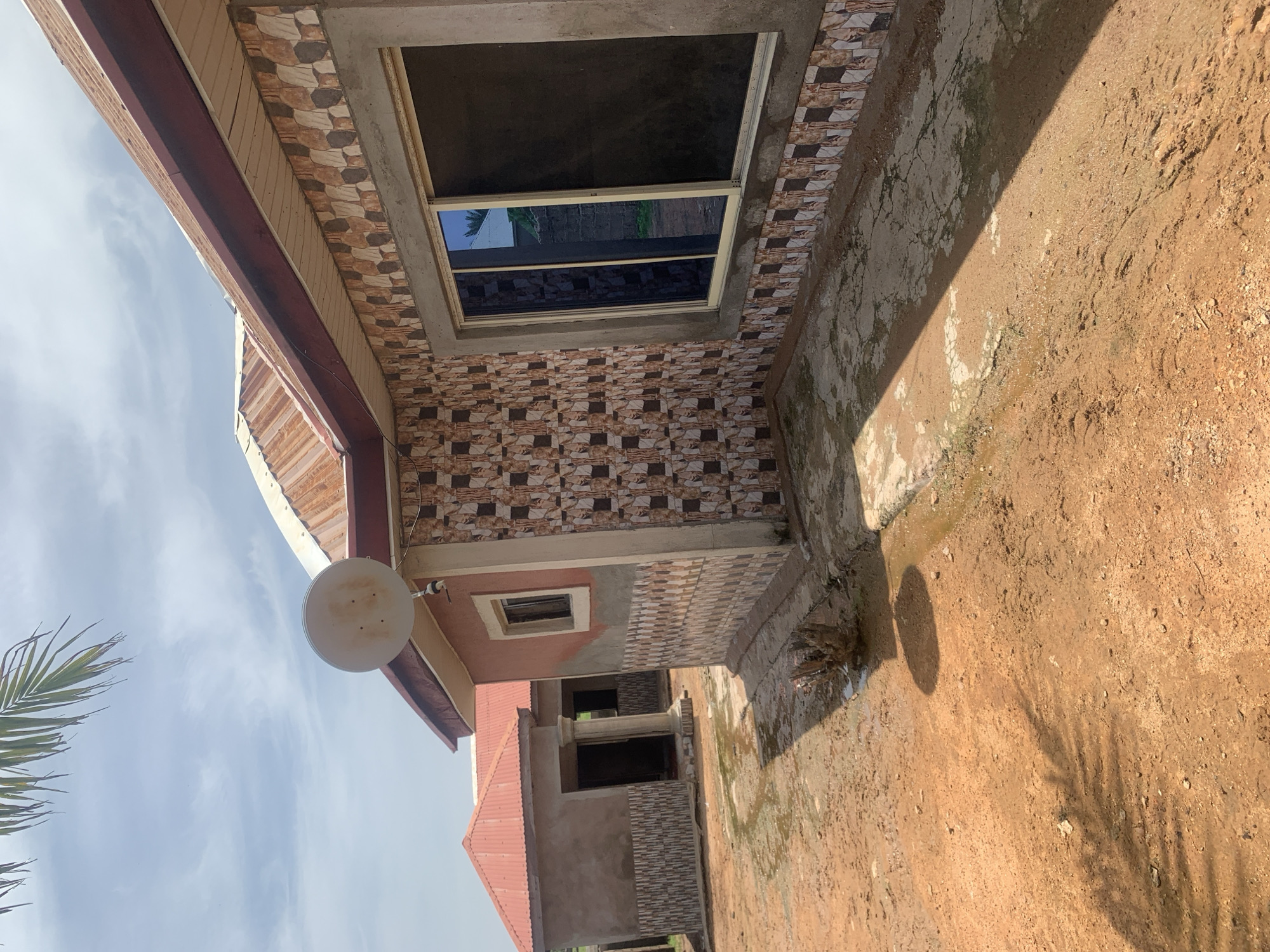 The image depicts an outdoor scene in Jauro Yinu, Nigeria, featuring two unfinished or newly constructed buildings. The foreground is dominated by reddish-brown soil, with patches of dried and wet mud, suggesting recent rain. Shadows of palm fronds fall across the ground. A small, dark, root-like object is visible in the mud near the right edge of the foreground.
The building on the right is partially tiled with a geometric pattern of brown, beige, and white tiles around its base and up to a window. The upper portion of this building is covered in a textured, light brown plaster. It has a large, modern sliding window with reflective blue-tinted glass. A satellite dish is mounted on the roofline, which is constructed of corrugated metal with a reddish-brown trim. The roof of this building has exposed metal beams and appears to be unfinished.
The building on the left is simpler in construction. It has a red corrugated metal roof and a plain, grey plastered exterior. A small window is visible on its left side. The front of this building features an open doorway with two columns flanking the entrance, and more of the patterned tile is visible at the base. The area between the two buildings is also composed of the reddish soil and shows signs of water accumulation. The overall environment appears to be rural or semi-developed, with a bright sky and scattered clouds.