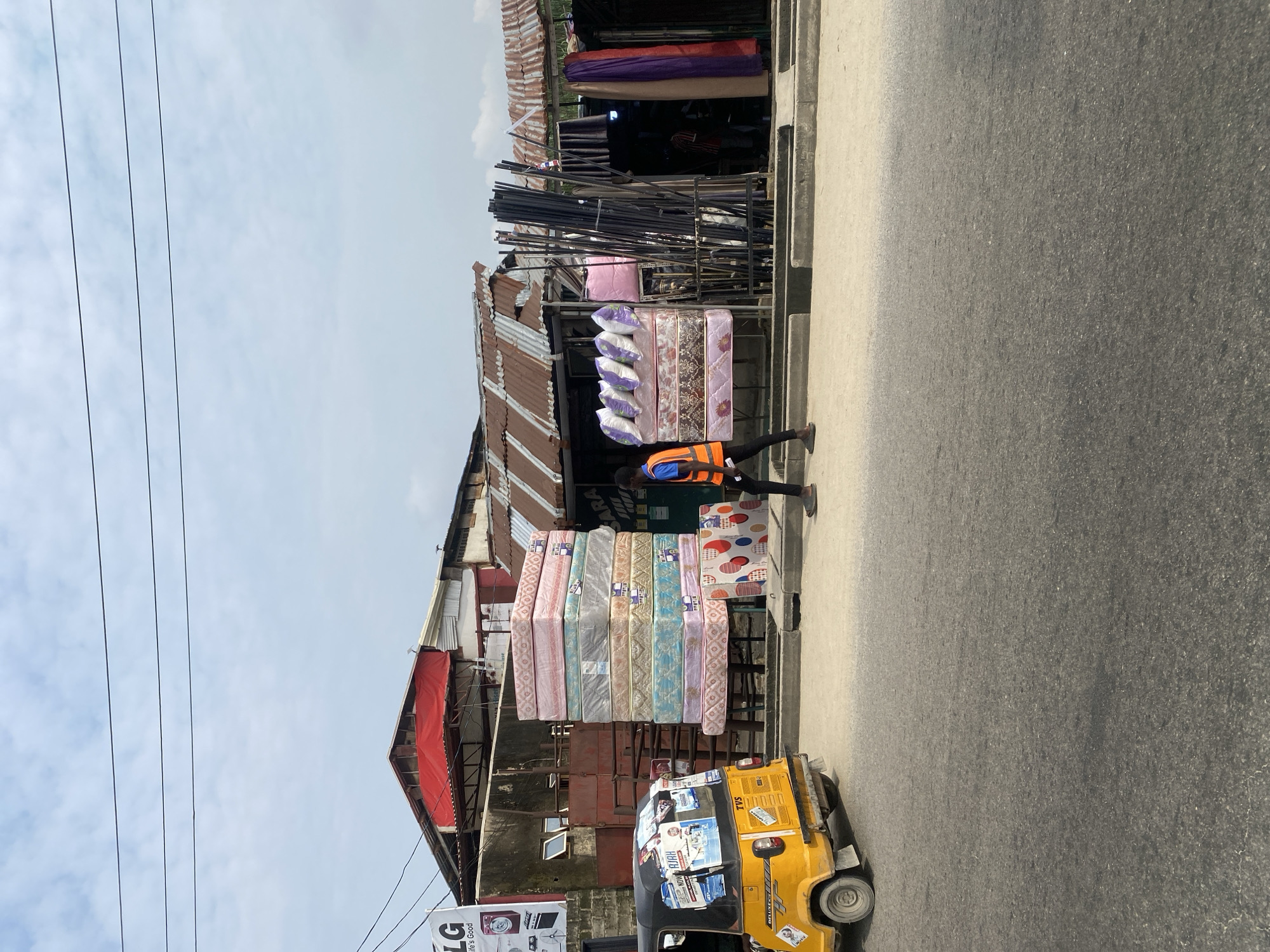 A street scene shows a yellow auto-rickshaw parked on the right side of the image, facing left, with posters adhered to its sides. A person wearing an orange vest walks to the right in front of a stack of mattresses. The mattresses are colorful and patterned, some wrapped in plastic. Behind the mattresses, there are structures with corrugated metal roofs, suggesting a market or shop area. In the background, buildings and electrical wires are visible against a cloudy sky.