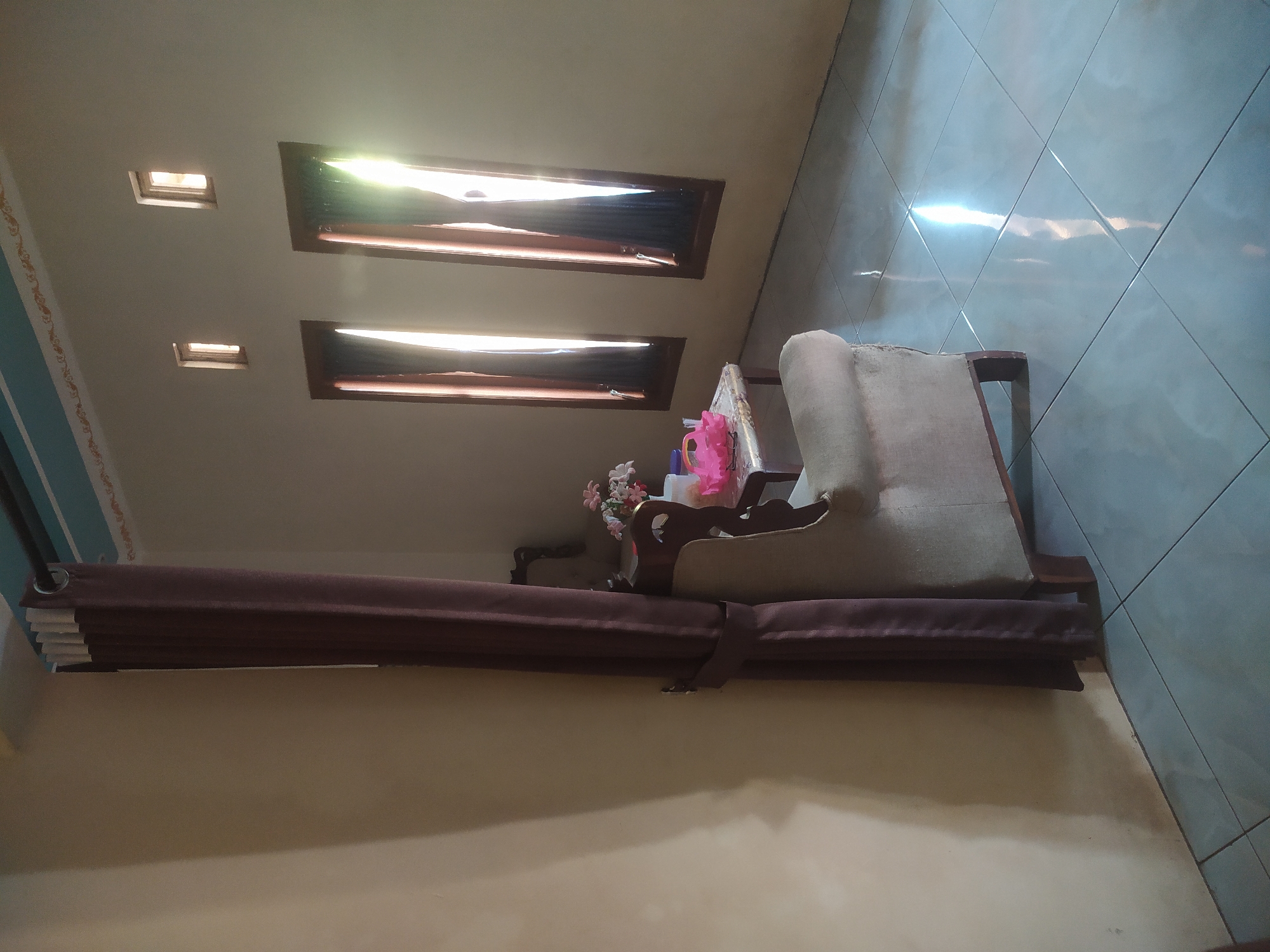 The image depicts an indoor scene, likely a living room or common area, with a tiled floor and light-colored walls. Sunlight streams in through two tall windows, illuminating the room and casting reflections on the floor. A dark brown curtain is partially drawn on the left side of the frame, obscuring a portion of the wall and a chair. To the right of the curtain, a light-colored armchair is visible, with its back and armrest facing the viewer. Beside the armchair, a small table holds a bouquet of flowers and a pink object, possibly a tissue box or a toy. The windows are framed with dark wood, and smaller, rectangular vents are present higher up on the wall. The ceiling features a decorative border. The overall impression is that of a well-lit, lived-in space. The location in Lopait, Indonesia, is not visually discernible from the image itself, but it provides context for the scene.