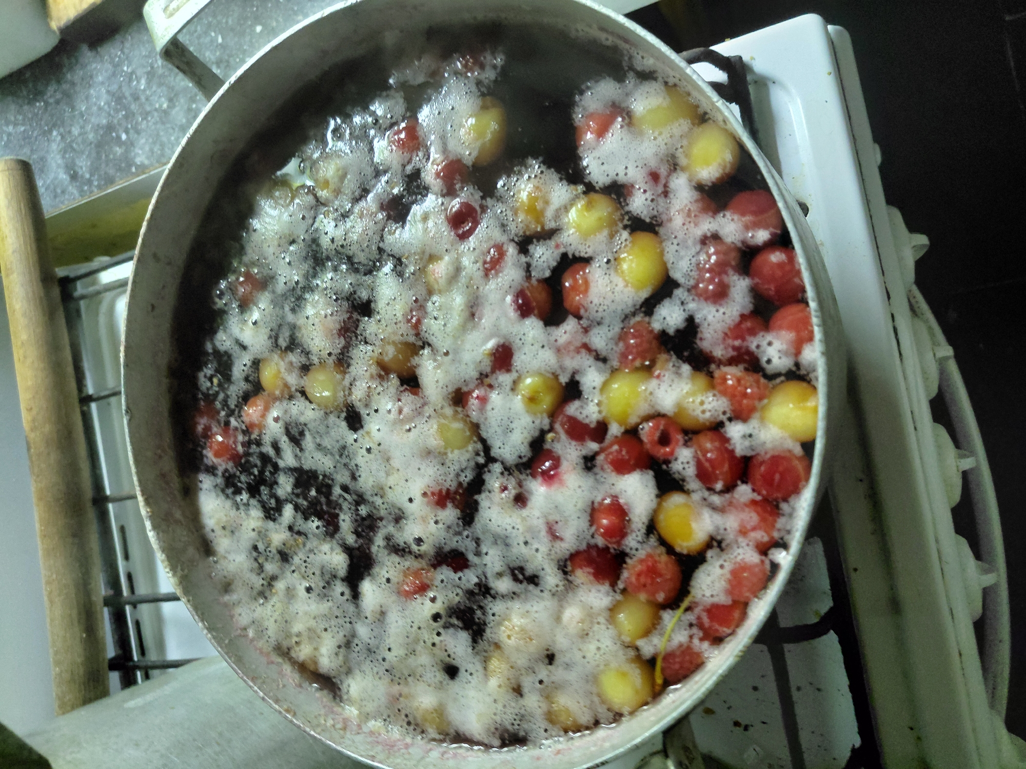 A close-up, overhead shot captures a pot of simmering fruit, likely cherries and possibly raspberries, bubbling with foam on a white gas stove. The fruits, a mix of deep red and pale yellow, are partially submerged in a liquid that has turned dark from their juices. The foam, white and airy, covers the surface, obscuring some of the fruit and creating a textured visual. The pot itself is a standard, slightly worn metal pot, its rim visible around the edges of the frame.
The setting appears to be a kitchen, indicated by the presence of the stove, which has a white enamel finish. To the left of the pot, a wooden object, possibly a utensil or part of the stove's design, is visible, adding a rustic touch. The background is dark and out of focus, suggesting an indoor environment. The lighting is not particularly strong, giving the image a somewhat subdued, perhaps even domestic, feel. There is no indication of the time of day or weather. The activity is clearly the cooking or preserving of fruit, likely for jam or compote. The overall impression is one of home cooking or food preparation. No text is visible in the image.
