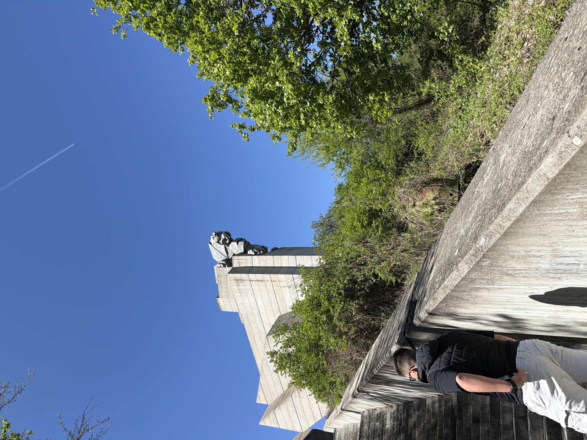 A low-angle shot captures the Founders of the Bulgarian State Monument in Shumen, Bulgaria, under a clear blue sky. The monument, predominantly white and angular, is partially obscured by greenery. A person wearing a black vest and light-colored pants stands on a gray, tiered concrete structure, possibly stairs, with one hand near their back pocket. A jet stream is visible in the upper left portion of the sky. Trees and foliage border the right edge of the image.