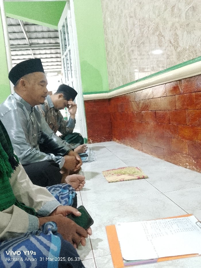 The scene takes place in a room with tiled floors and brick walls. The room appears to be part of a home or a small community building. There are four men sitting on the floor. One man is wearing a blue shirt, black pants, and a black cap. He is seated at the front of the image, looking at his phone. The second man is wearing a grey shirt, black pants, and a black cap. He is seated behind the first man, looking down at a small book. The third man is wearing a green shirt, black pants, and a black cap. He is seated behind the second man, looking down at a small book. The fourth man, partially obscured by the first man, appears to be wearing a dark blue shirt. They are all sitting together, possibly engaging in conversation or a religious study session. The room also contains two notebooks on the floor in front of them. The location of this scene is Anjatan, Indonesia, at coordinates -6.419 latitude and 107.979 longitude.