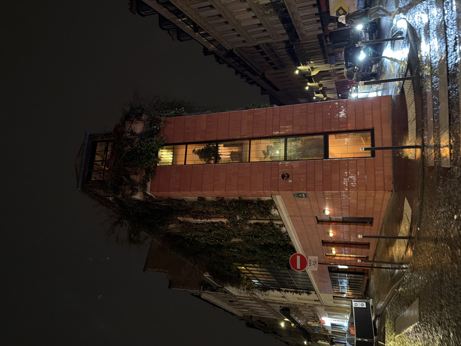 A brick building at night is covered in ivy. The large, rectangular windows are lit from within, revealing houseplants. Reflections from the rain-slicked street are visible. A "no bicycles" sign is seen, along with street lamps. The sky is dark, and other buildings can be seen in the background, suggesting a city location.