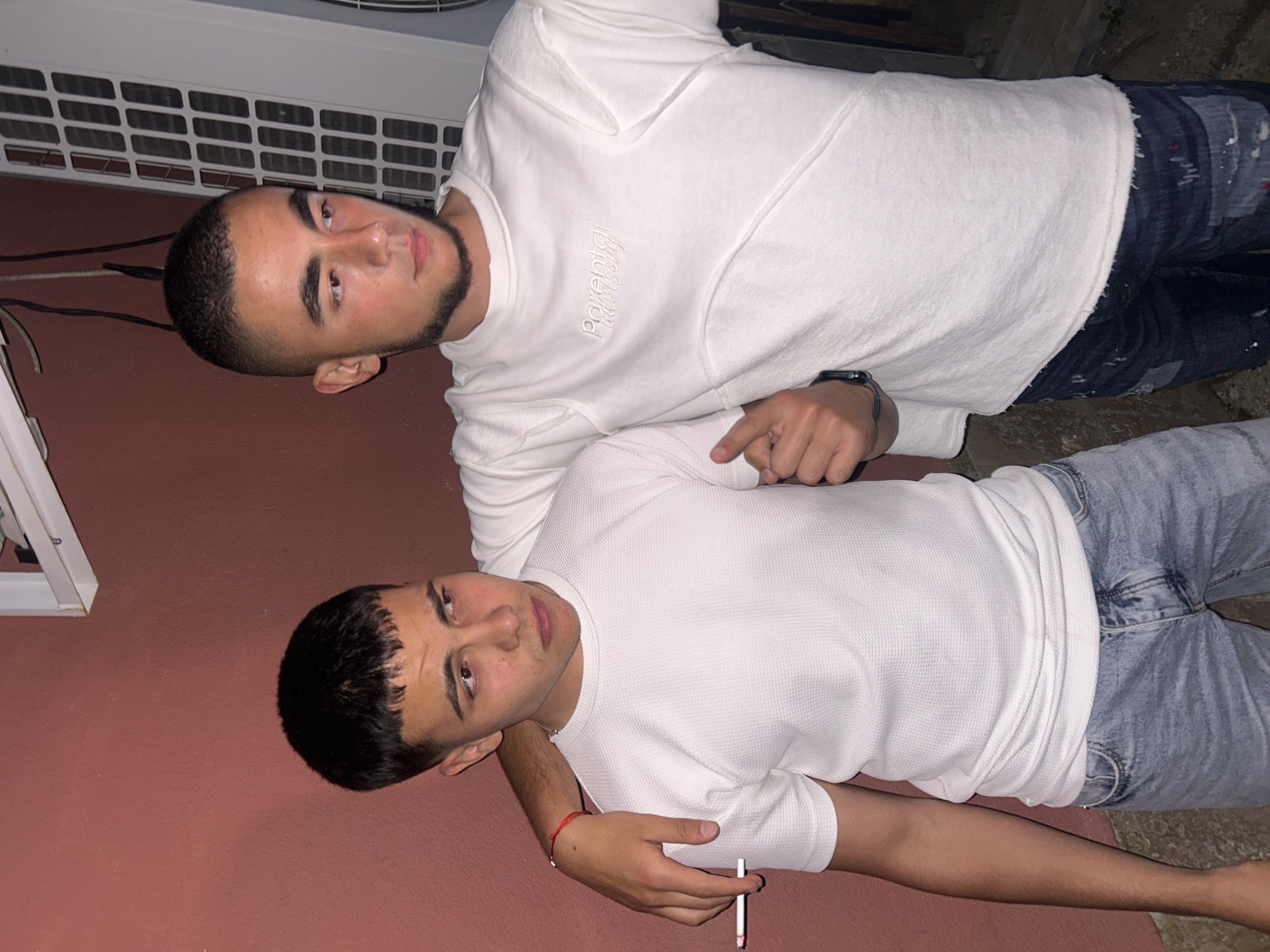 Two young men are lying on what appears to be an outdoor surface. The man on top wears a white sweatshirt with the text "Parental Philosophy" written on it, along with dark denim jeans. He also wears a watch on his left wrist and points with his right index finger. The man below wears a white waffle knit t-shirt and light blue jeans. He holds what looks like a cigarette in his left hand, while his right arm is around the first man's shoulder. The environment suggests an urban setting. There is no specific indication of Elhovo, Bulgaria. The background includes a red-painted wall, a white air conditioning unit, and other indistinct objects.
