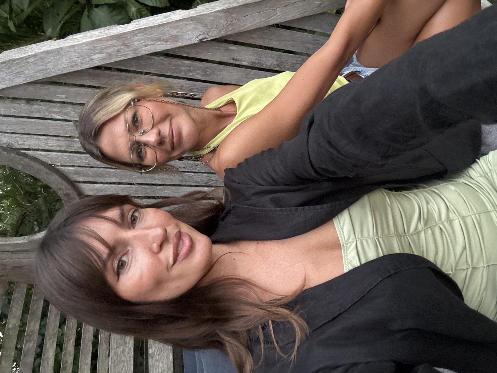 Two women are sitting on a wooden bench outdoors, likely in a garden setting. The woman on the left has long, dark brown hair and is wearing a black blazer over a light green ruched top. She is looking towards the camera with a subtle smile and has a nose ring. The woman on the right has blonde hair, wears large glasses, and is dressed in a light green halter top and denim shorts. She is also looking towards the camera. The background consists of wooden slats and lush greenery. The image appears to be a selfie taken by one of the women.