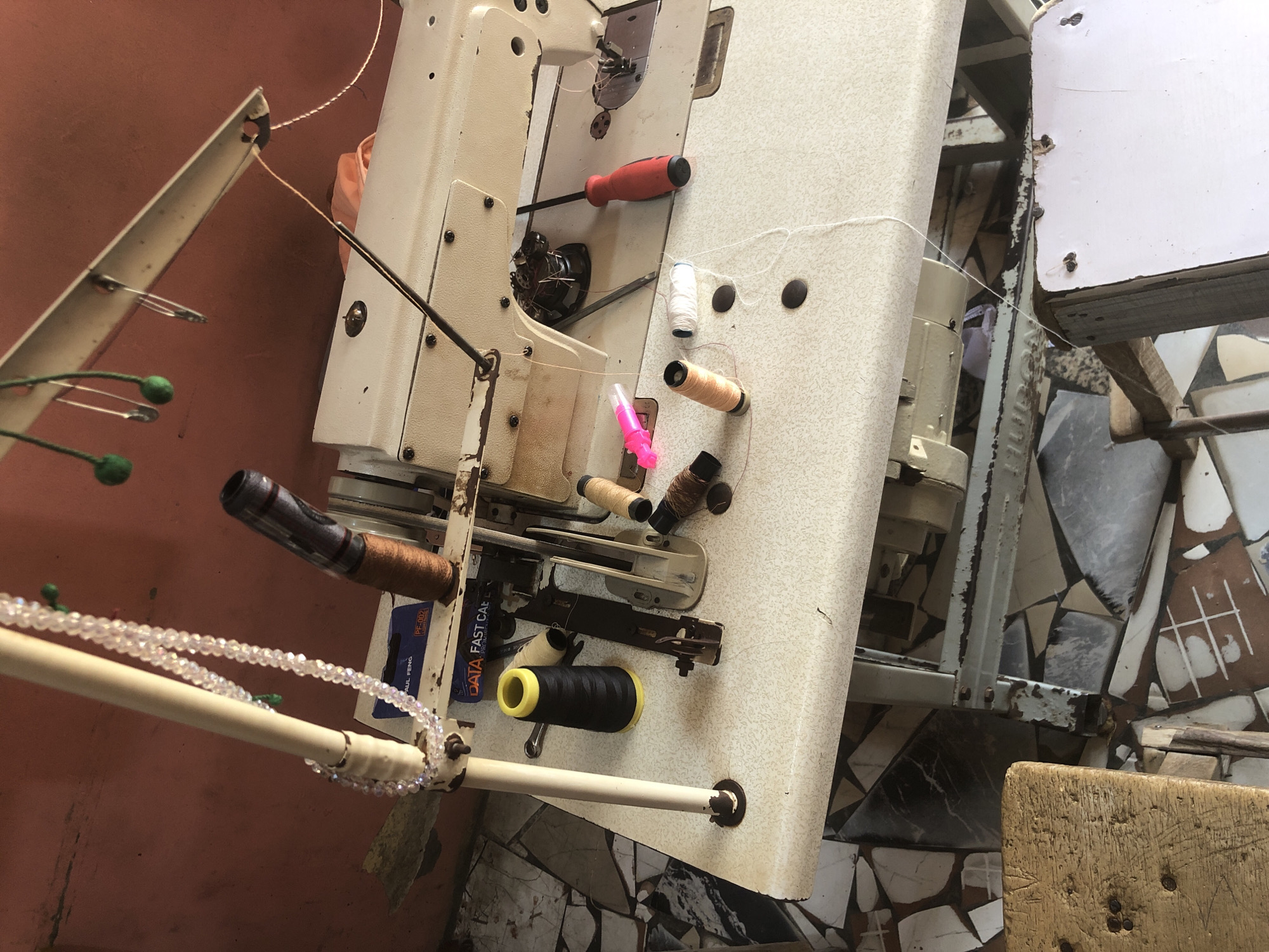 This image depicts a sewing machine and various sewing supplies, likely in a workshop or home setting in Ado, Nigeria. The sewing machine is cream-colored with a visible needle and presser foot. Spools of thread in black, white, and beige are scattered on the machine's surface. A red-handled screwdriver rests near the machine, along with a pair of scissors and a pink plastic bobbin holder. A beaded necklace hangs from a white pole to the left of the machine, and some green felt balls are suspended from above. The background shows a reddish-brown textured wall. The floor beneath is a mosaic of broken tiles in various shades of brown, white, and grey. A wooden stool is partially visible in the lower left corner. The scene suggests an active sewing area with materials readily accessible for use.