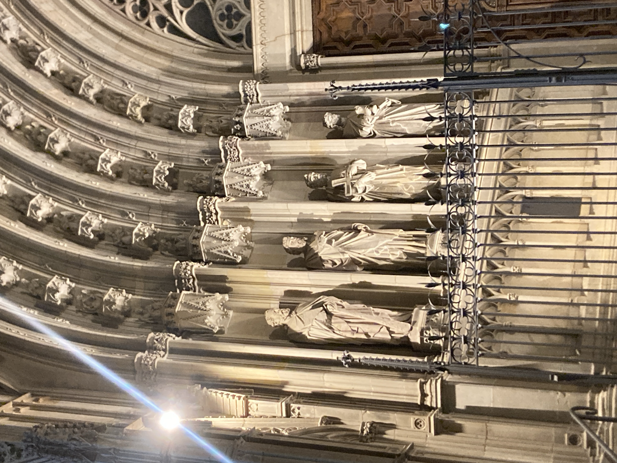 The image captures the facade of a Gothic-style building in Barcelona, Spain, at night. Five stone statues of male figures, dressed in robes and appearing to be historical or religious figures, are displayed in arched alcoves. The statues are illuminated by artificial light, casting shadows and highlighting the architectural details of the building, which include ornate carvings, columns, and vaulted ceilings. A decorative metal fence is positioned in front of the statues, obscuring a direct view of their bases. The overall scene suggests a cultural or religious landmark, likely a church or cathedral, given the architectural style and the presence of the statues.