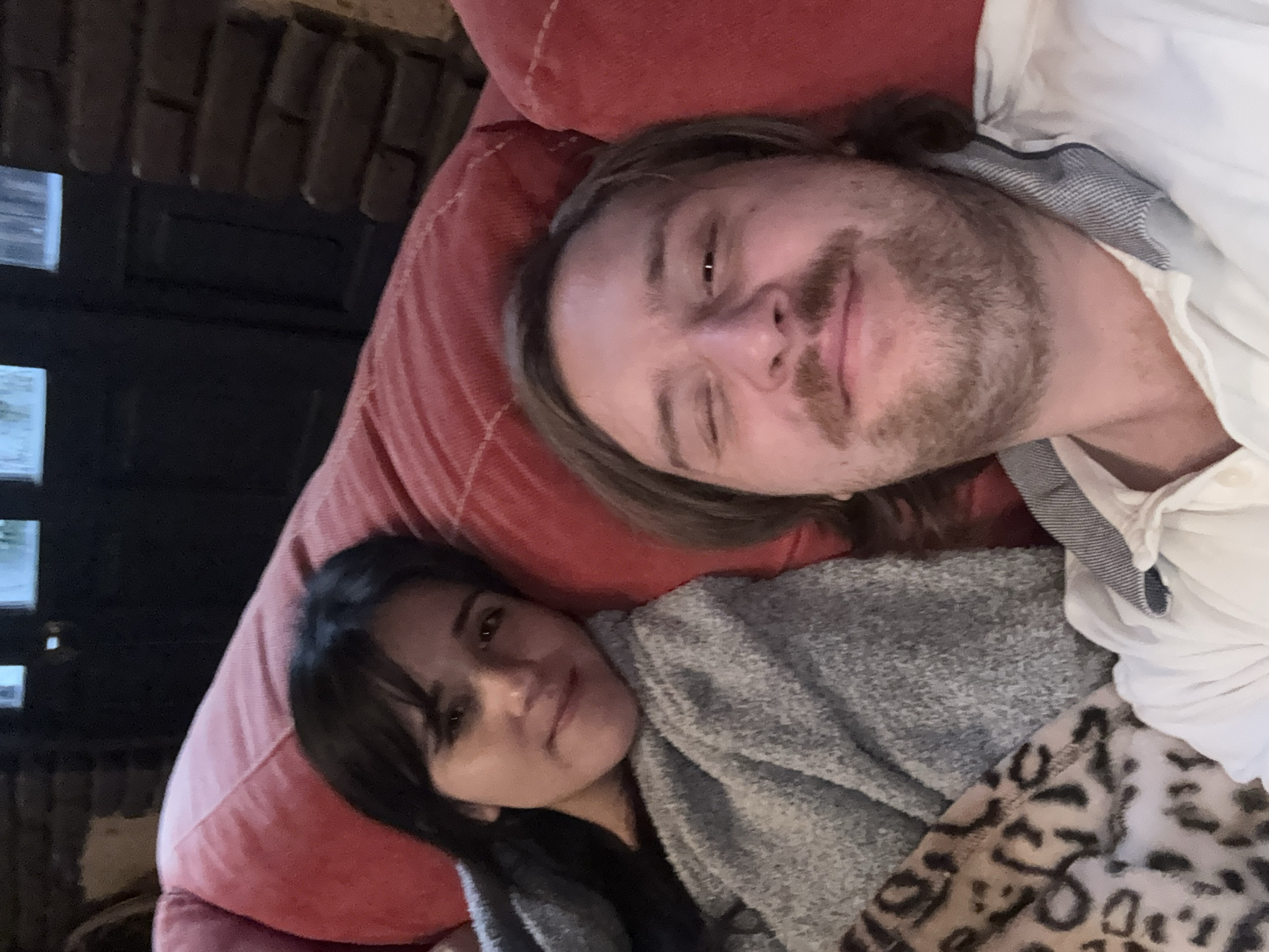 A man and a woman are lying on a red couch. The woman is partially covered with a gray blanket and an animal print blanket. The man is wearing a white shirt. The location appears to be indoors, with a window and textured wall visible. It is not possible to confirm that the location is in the city of Cañada de Madero, Mexico from the image provided.