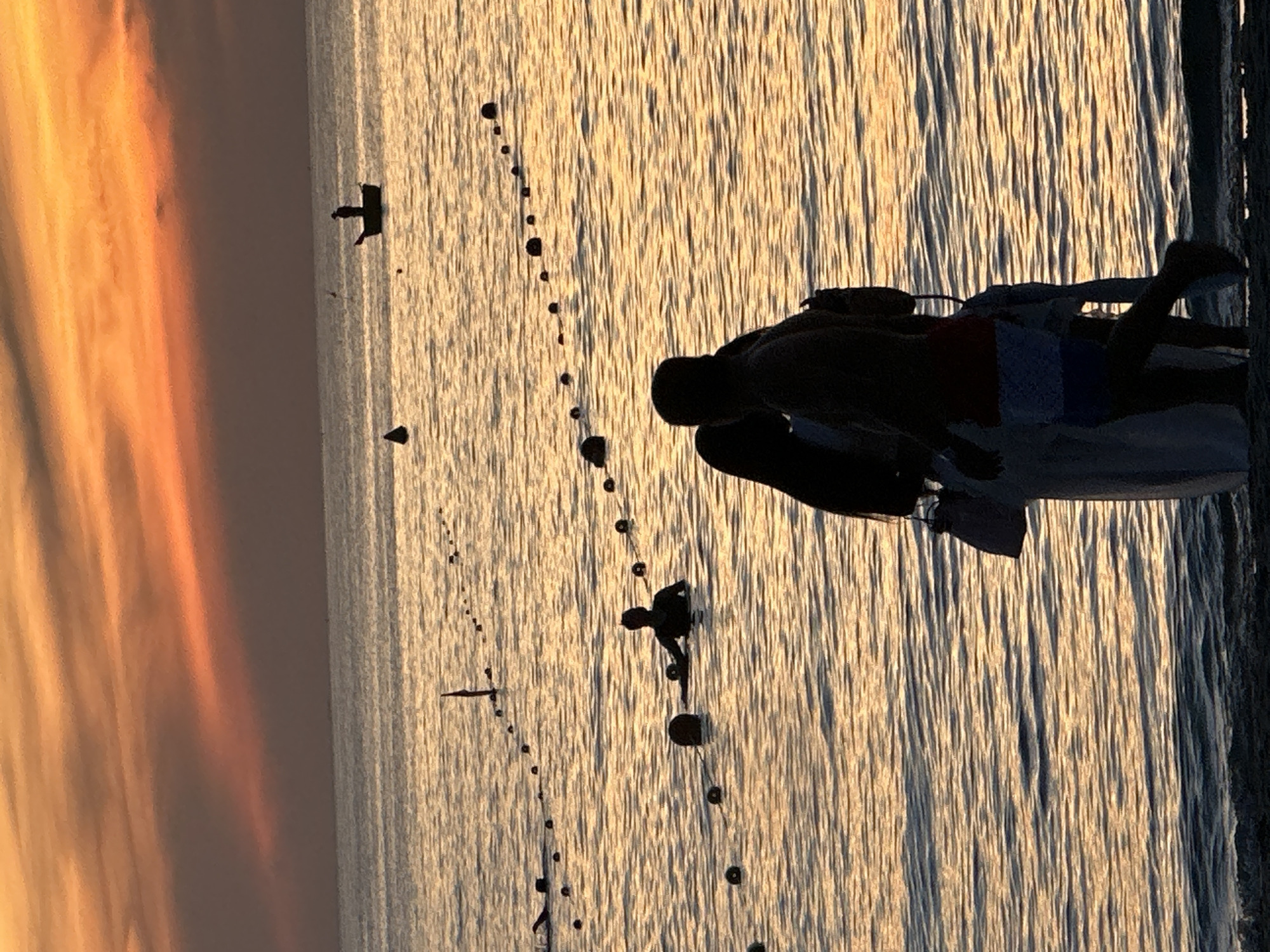 The image captures a sunset scene at the beach in Đà Nẵng, Vietnam. The sky is illuminated with a warm orange and yellow gradient from the setting sun, casting a golden glow on the rippling water. In the foreground, silhouetted figures of people are visible. Two individuals, a man and a woman, stand close together on the sand, facing away from the camera. The man appears to be wearing shorts, and the woman is holding a bag. Further out in the water, several other silhouetted figures can be seen. One person is swimming, while another is standing in the water. A small boat with a single person in it is visible in the distance, contributing to the tranquil and serene atmosphere of the coastal setting. A rope with buoys stretches across the water, marking a section of the beach.