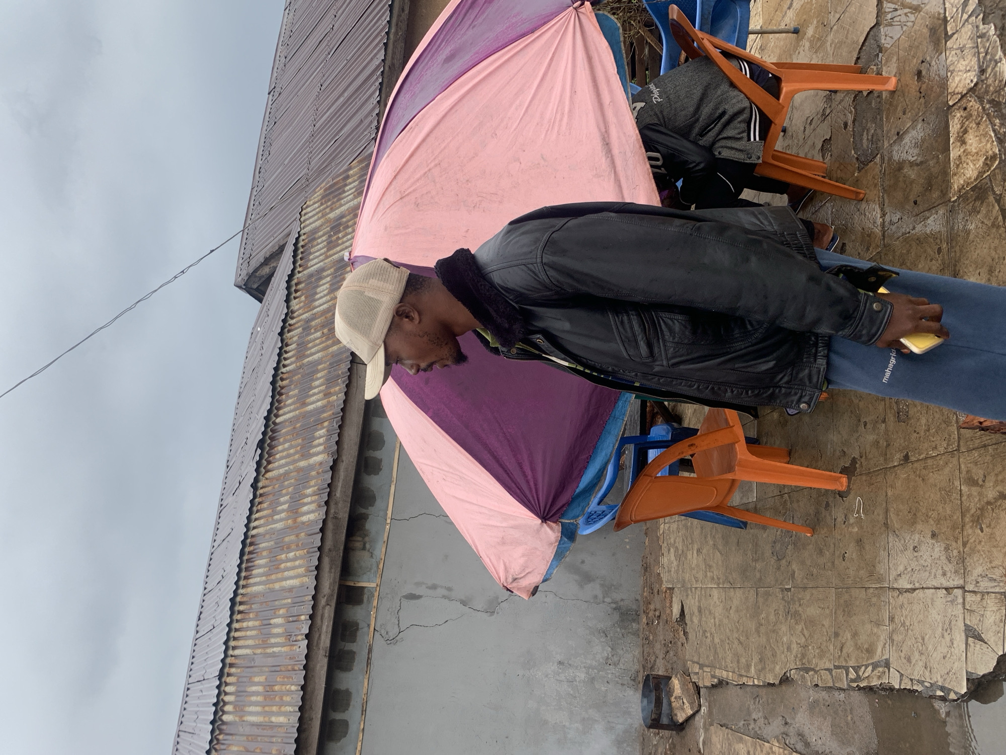 A man wearing a beige baseball cap and a black jacket stands in front of a large, open umbrella with pink and purple panels, in what appears to be an outdoor seating area. He is facing to the left, with his right hand holding a yellow smartphone. To his right, partially obscured by the umbrella, is another person sitting on an orange chair. The ground is paved with square tiles, some of which are wet and cracked. Behind the umbrella, there are blue and orange plastic chairs. A metal brazier with some wood is visible to the left of the man. In the background, a corrugated metal roof is attached to a grey, cracked wall. The sky is overcast and grey. This scene is likely from Jos, Nigeria, given the presence of corrugated iron roofing and outdoor seating arrangements common in such environments.