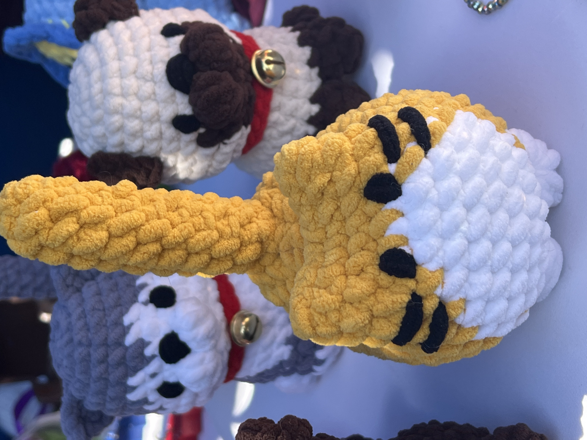 The image shows a close-up of several handmade crocheted toys. The toys appear to be animal-themed, with visible examples including what appear to be a tiger or lion (yellow and white with black stripes), a cow (white with brown patches), and a wolf or husky (gray and white with black markings). The toys are made from soft, textured yarn and have details like collars with bells. The background is blurred, suggesting a shallow depth of field. The toys are likely displayed on a light-colored surface, possibly a table or display stand. There is no information about the location in Arica, Chile, to include in the description.