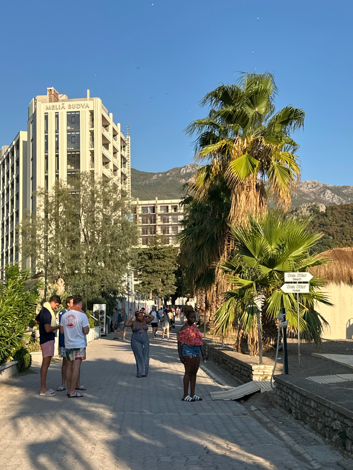 In the city of Rafailovići, Montenegro, an outdoor scene unfolds under a clear blue sky. A multi-story hotel, identified by the signage "MELIA BUDVA," stands prominently on the left side of the frame, its facade a light cream color with numerous balconies and dark-tinted windows. In front of the hotel, a paved walkway is populated by several individuals.
A group of three young men are conversing near the left edge of the walkway. One, wearing a dark blue t-shirt and pink shorts, has a yellow towel draped over his shoulder. Another, in a white t-shirt with red lettering and patterned shorts, is facing away from the camera. The third, partially obscured, appears to be in a similar casual attire.
Further down the walkway, a woman in a striped blue and white halter top and matching wide-leg pants is posing, her hands on her head. To her right, closer to the center of the frame, another woman in a pink patterned tie-front top and denim shorts stands with her arms at her sides.
Several tall palm trees with lush green fronds dominate the right side of the image, casting shadows on the walkway. One palm tree has a directional signpost attached, with two arrows. The upper arrow points to "SEA STAR Beach," and the lower arrow points to "SEA STAR Hotel." In the background, behind the palm trees, is a building with a thatched roof, and beyond that, a range of rocky, tree-covered mountains. The ground on the right side of the walkway is covered in sand and gravel. The overall atmosphere suggests a warm, sunny day in a coastal resort town.