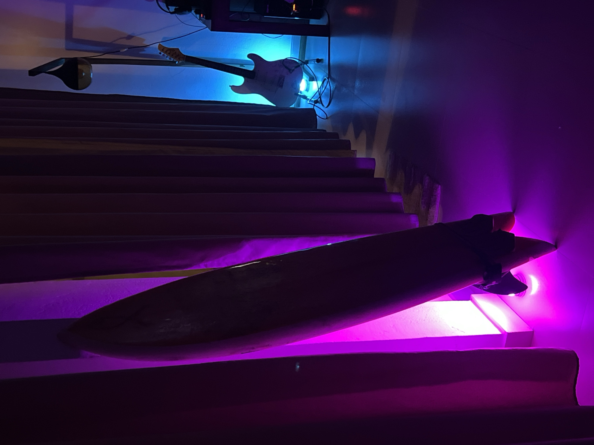 A staircase in Goviyapana, Sri Lanka, is illuminated with blue and purple lighting. A white electric guitar hangs on the wall above the stairs. A surfboard lies diagonally across the lower steps.
