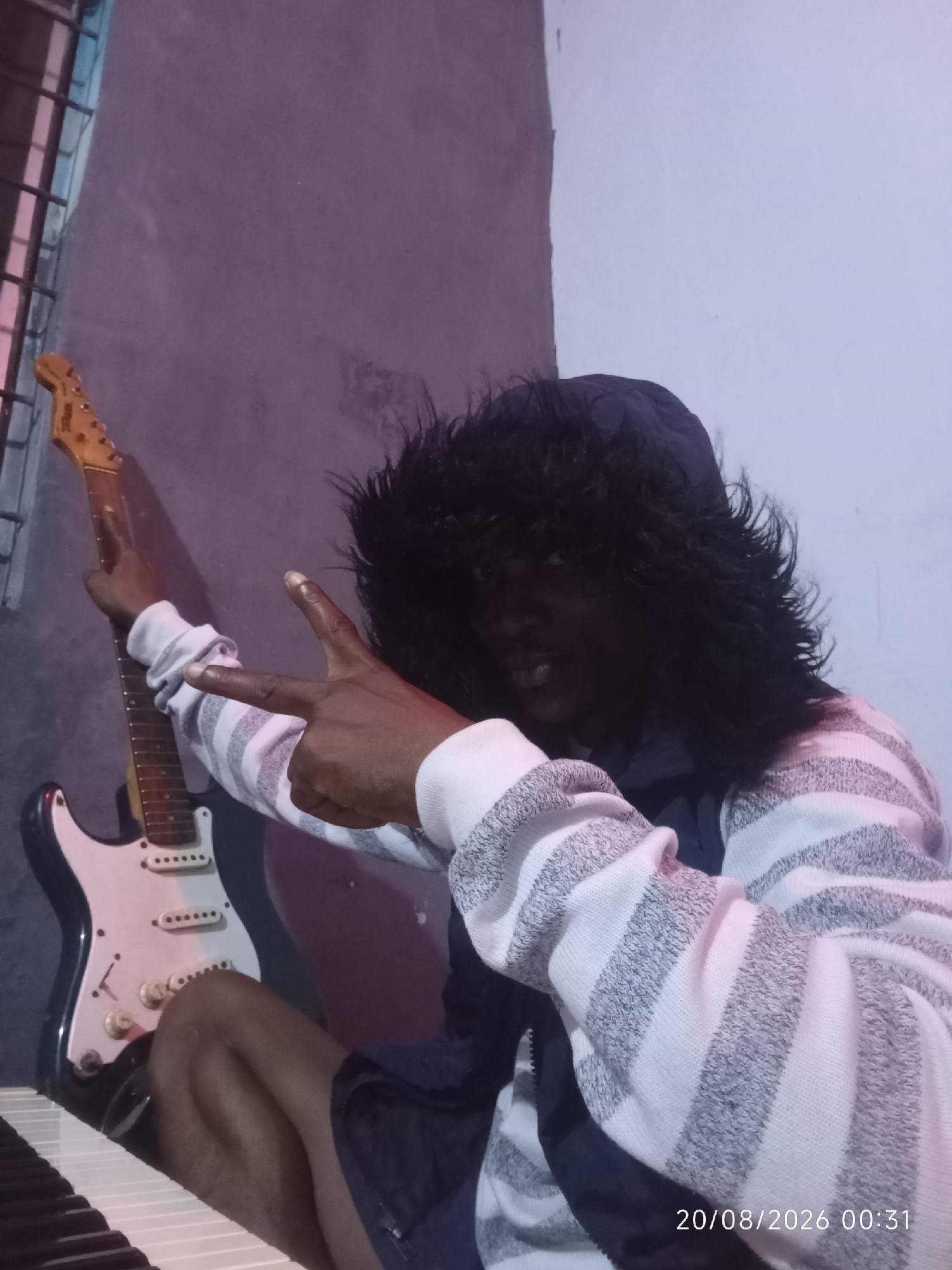 In Adankolo, Nigeria, an individual with dark skin and a dark, shaggy wig is seated, partially visible, in front of a white electric guitar and a keyboard. The person is wearing a blue hooded jacket and a white and grey striped sweater. Their right hand is raised, with the index and middle fingers extended in a peace sign gesture, while the left hand is also positioned in a similar gesture. The background consists of a textured wall. A timestamp, "20/08/2026 00:31", is visible in the lower right corner of the image.